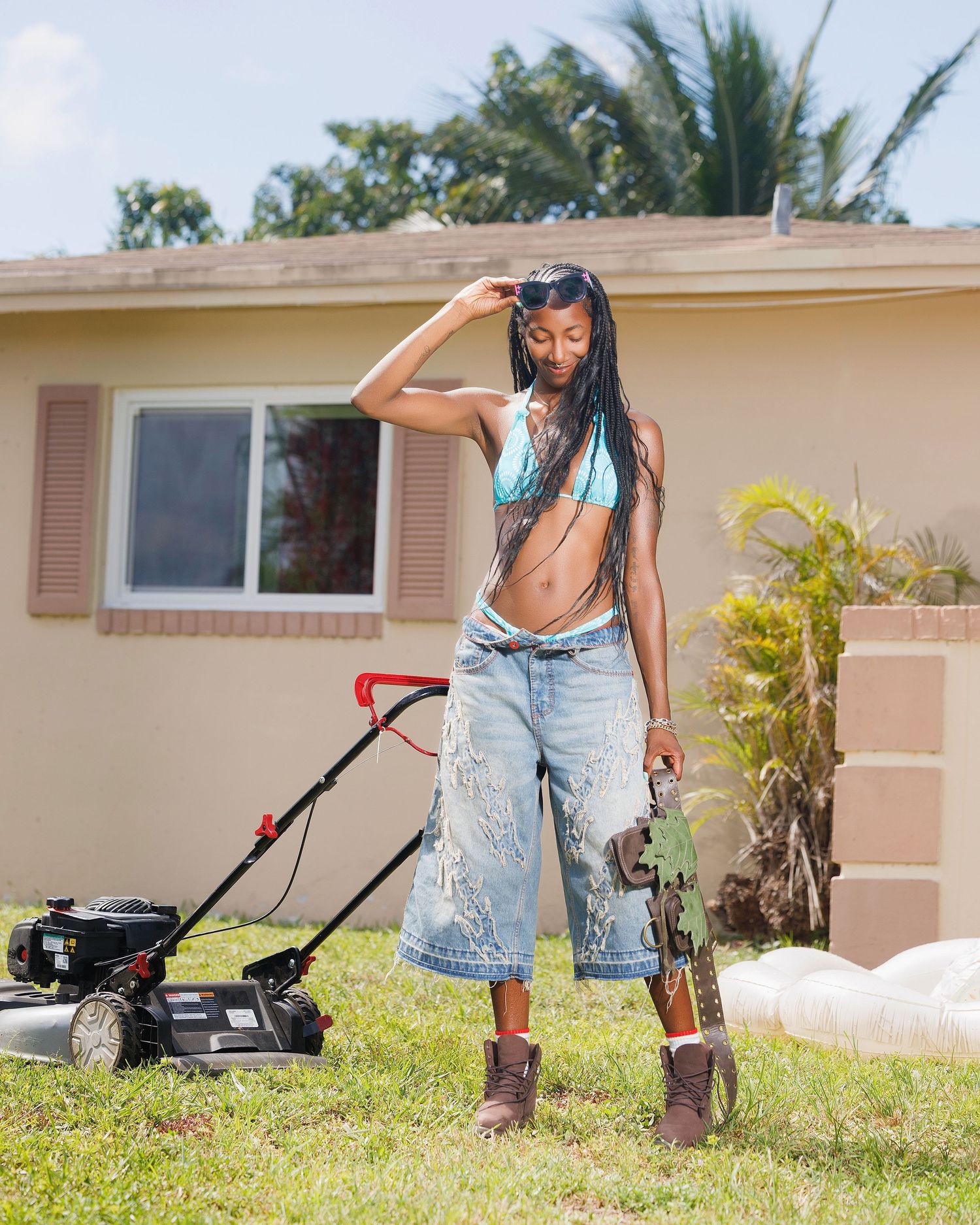 Woman in crop top and wide-leg jeans mowing lawn outside suburban home on sunny day.