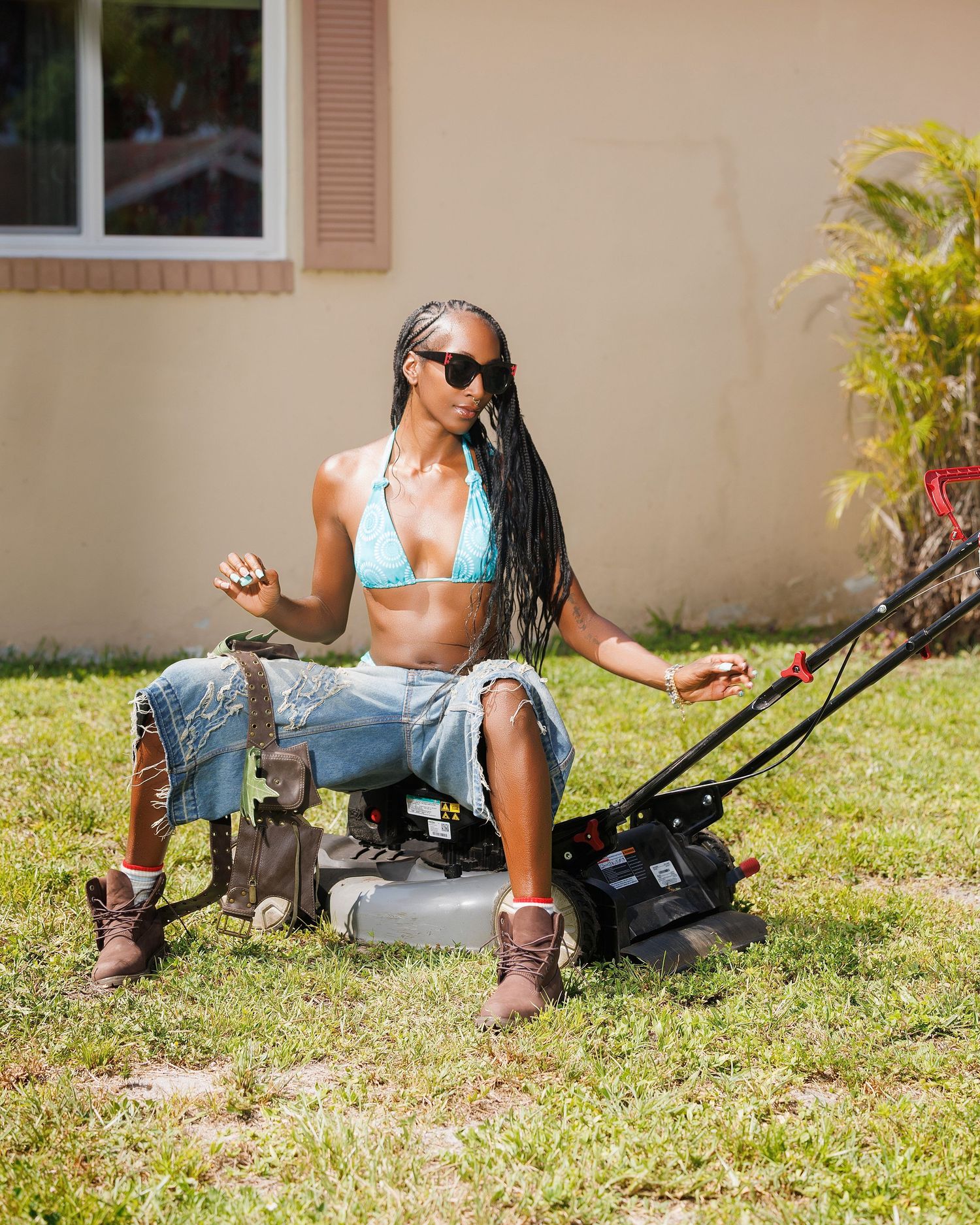 Woman in teal bikini top and denim shorts sitting on lawn mower in sunny yard wearing sunglasses and boots.