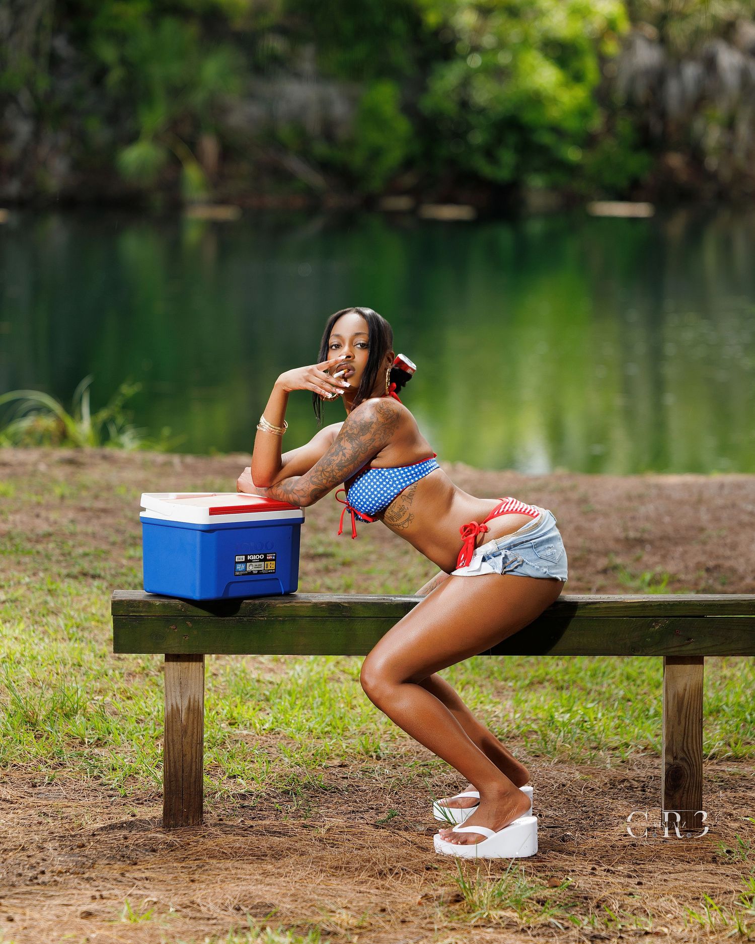 Woman in patriotic bikini top and denim shorts posing on picnic bench near blue cooler by scenic lake.