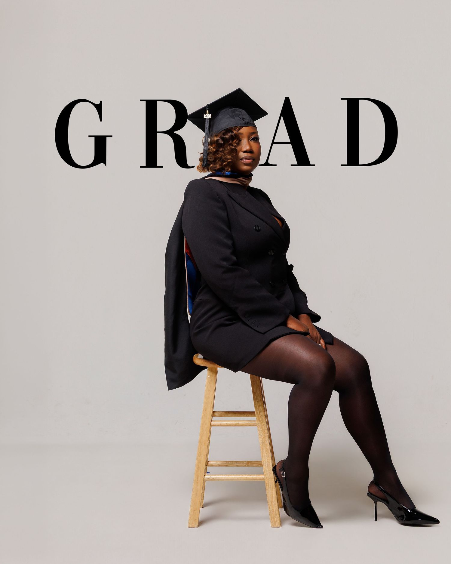 A graduate in cap and gown sits on a stool, posing confidently in front of a bold GRAD text backdrop.