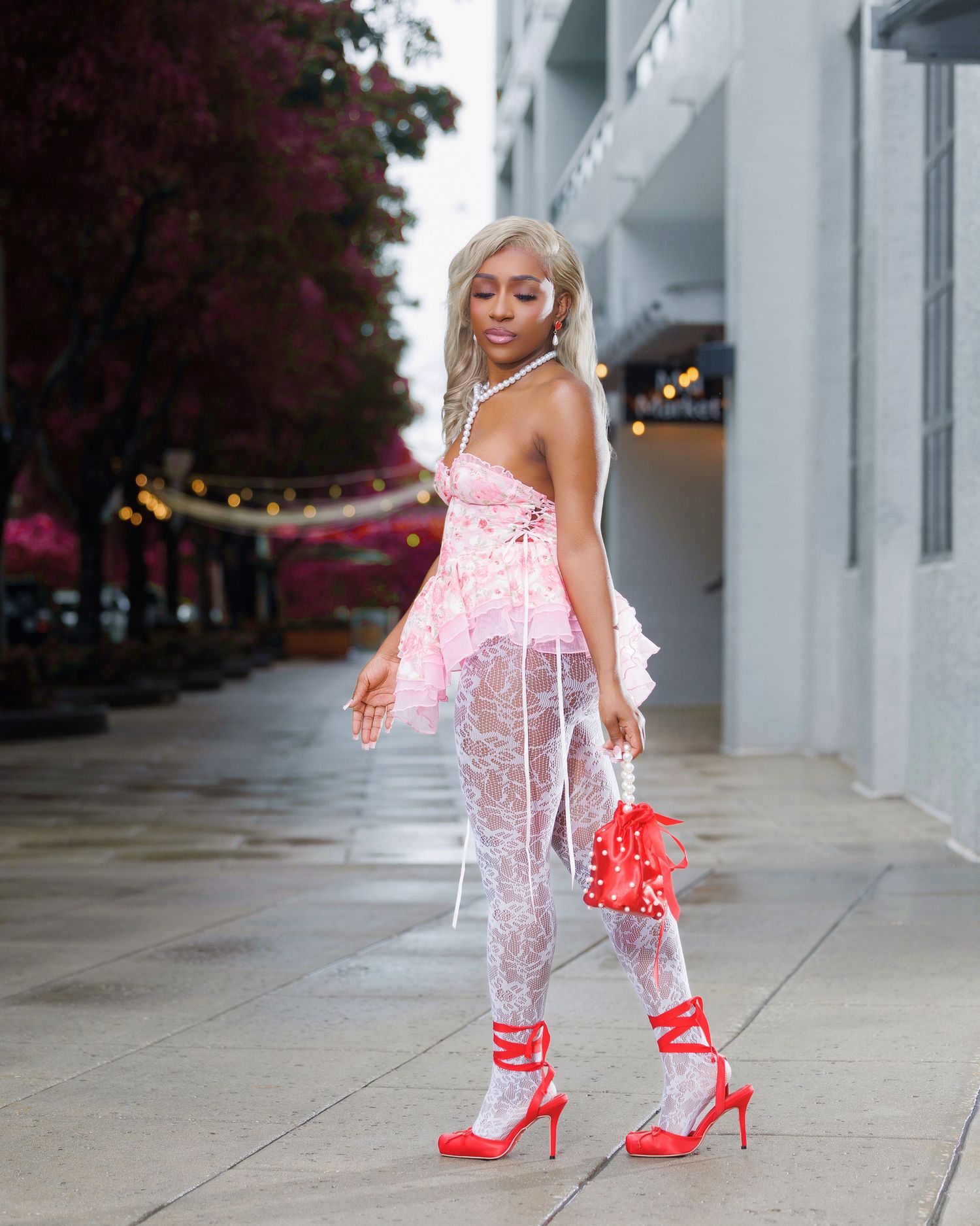 Woman in sheer lace dress with pink accents, white stockings, and red heels poses on urban sidewalk.