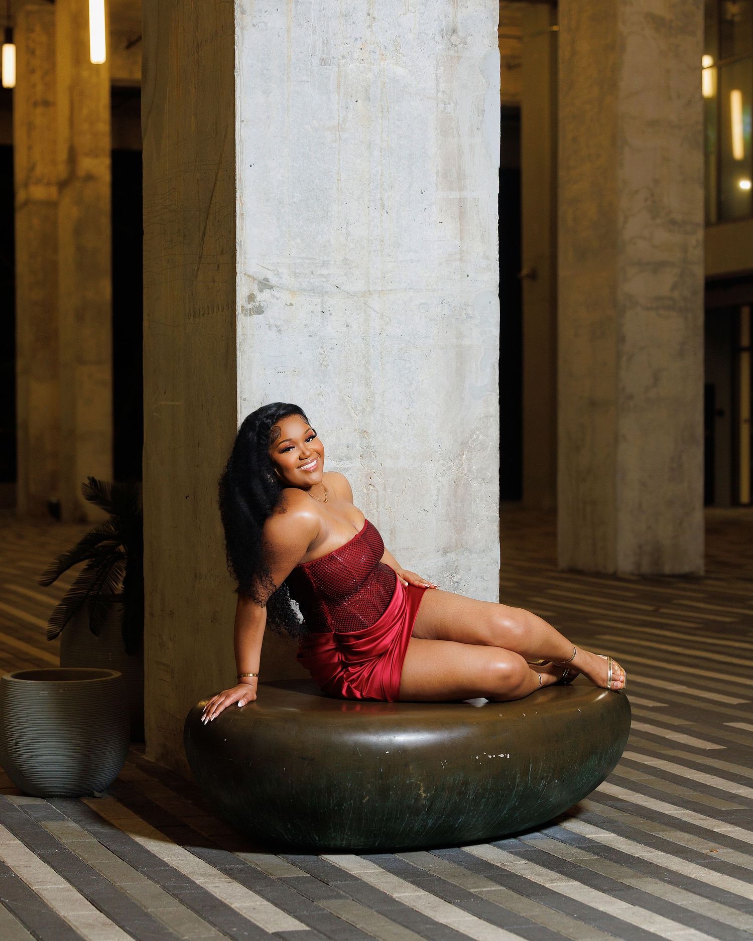 Woman in red satin dress posing on circular ottoman beside large concrete column in elegant interior.