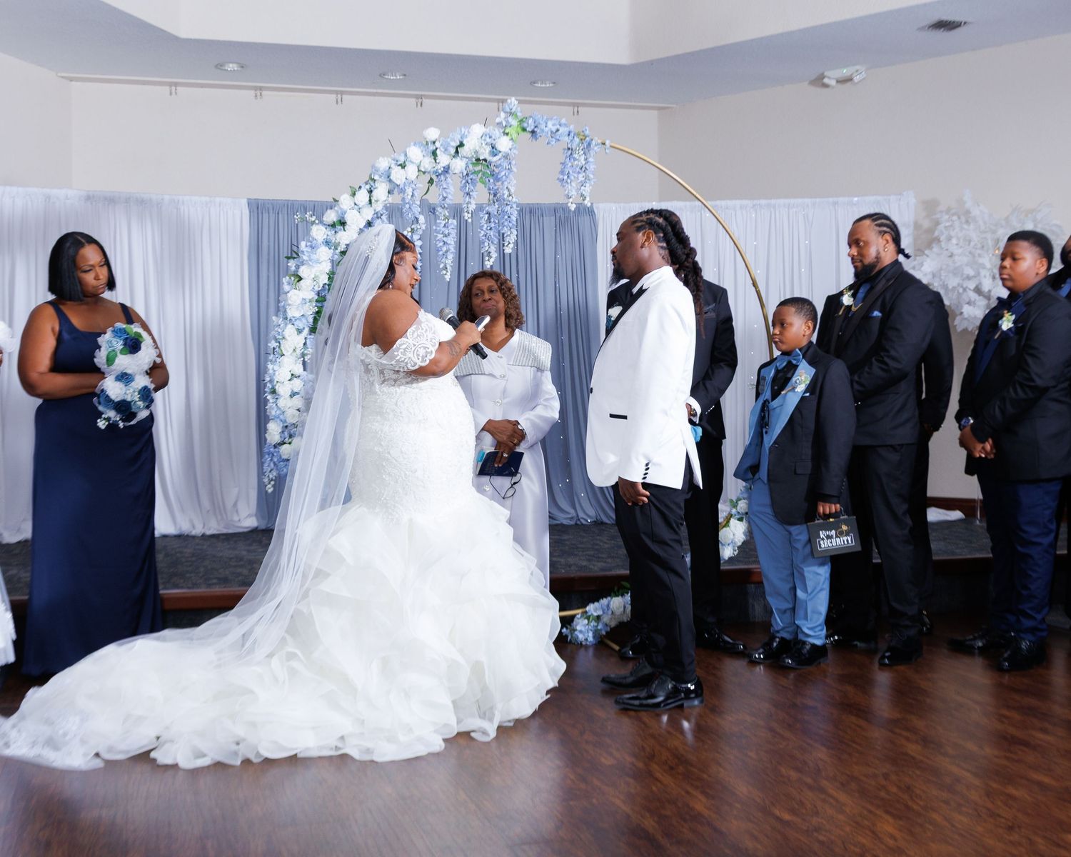 Bride and groom exchange vows at elegant wedding ceremony with blue floral arch and wedding party in navy attire.