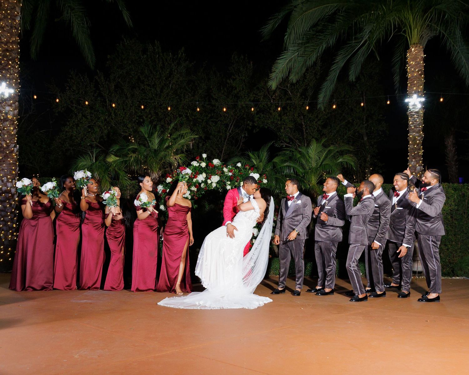 Wedding party poses outdoors at night, bridesmaids in burgundy gowns, groomsmen in gray suits, groom lifts bride.