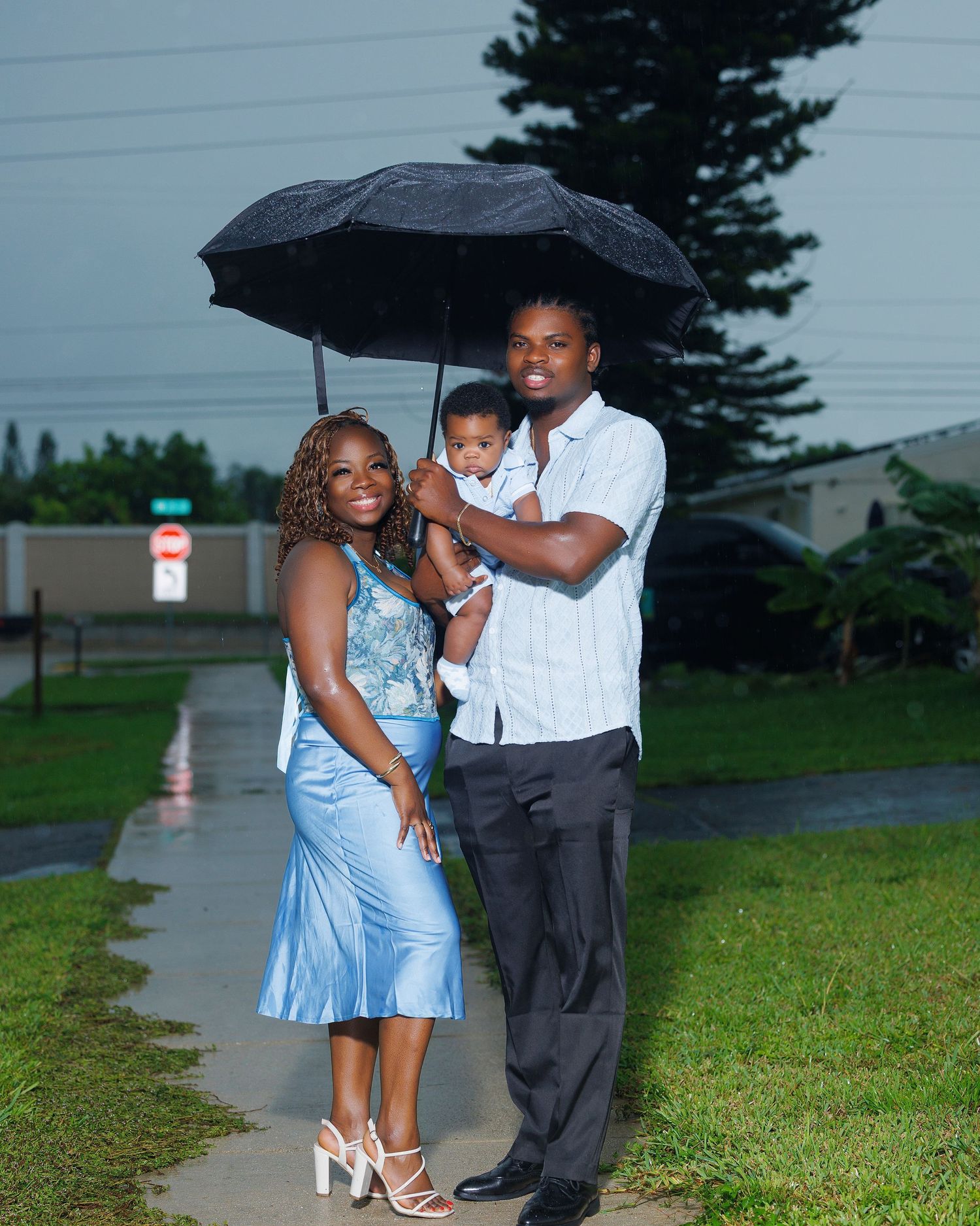 A family of three poses under a black umbrella in the rain, with parents holding their baby on a green lawn.