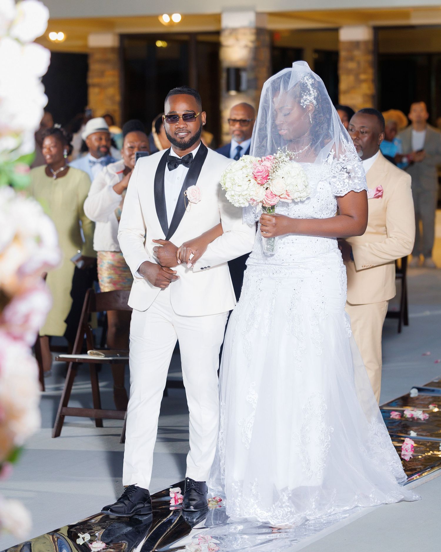 Groom in white tuxedo escorts bride in lace wedding dress down the aisle at an outdoor wedding ceremony.