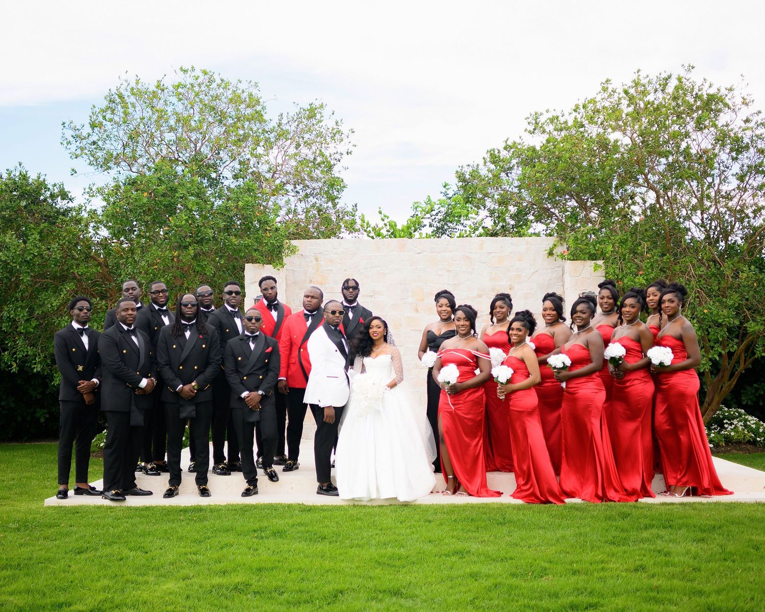 Wedding party poses outdoors, groomsmen in black tuxedos and bridesmaids in red gowns flank the bride and groom.