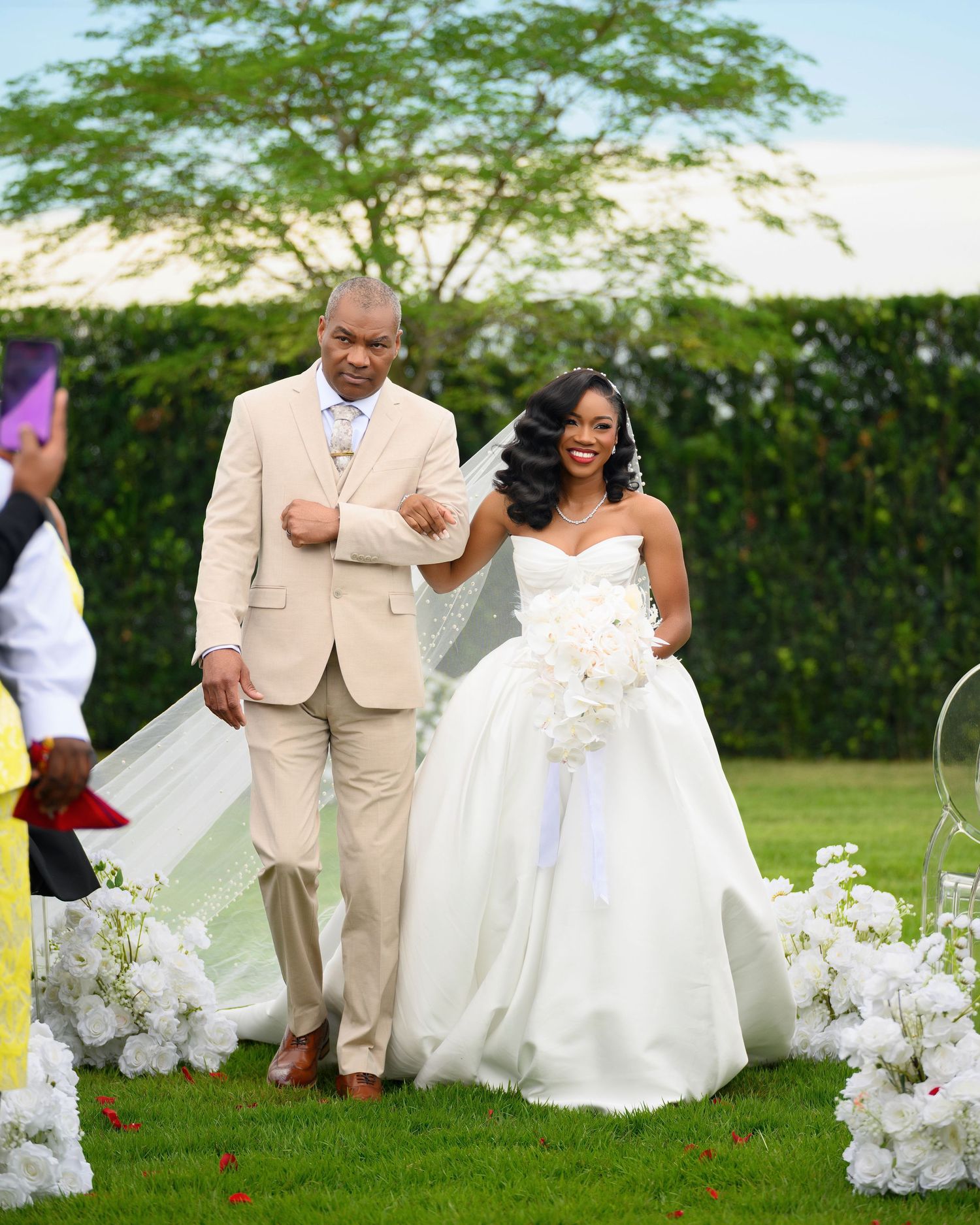 Bride in white ball gown walks down outdoor aisle with escort in tan suit, surrounded by white floral arrangements.