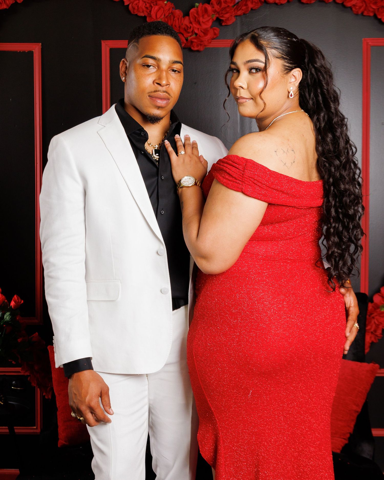Couple posing formally, man in white suit and woman in red off-shoulder gown, with red roses backdrop.