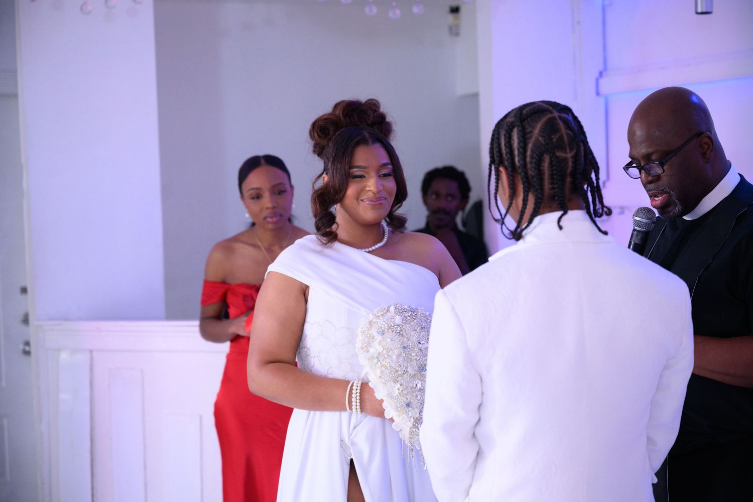 Bride and groom exchange vows at wedding ceremony with bridesmaid in red dress and officiant standing nearby.