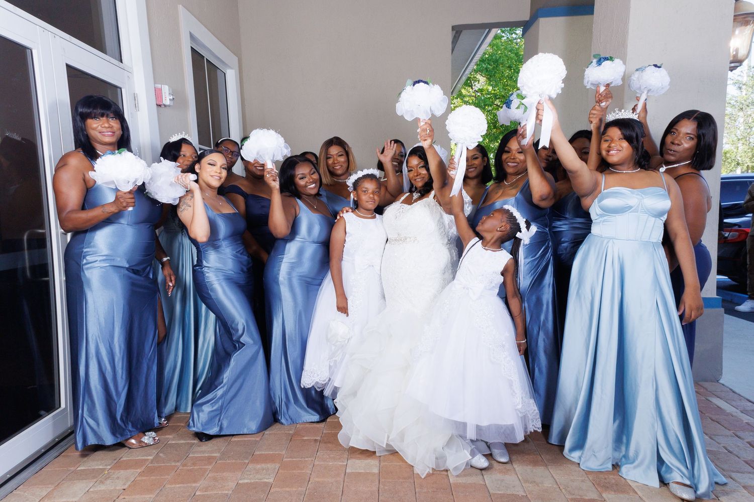 Bride in white gown with bridesmaids in blue satin dresses and flower girls holding white bouquets at wedding.