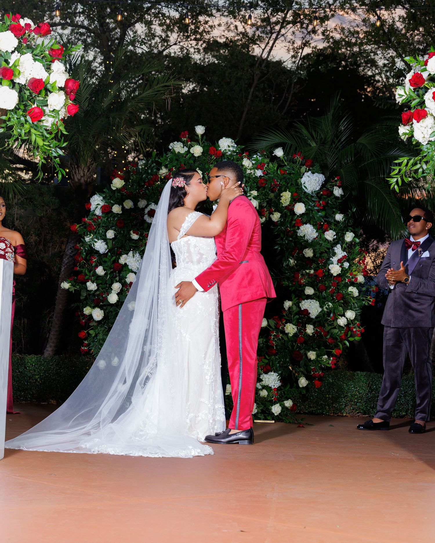 Bride and groom share first kiss at outdoor wedding ceremony before red and white floral arch backdrop.