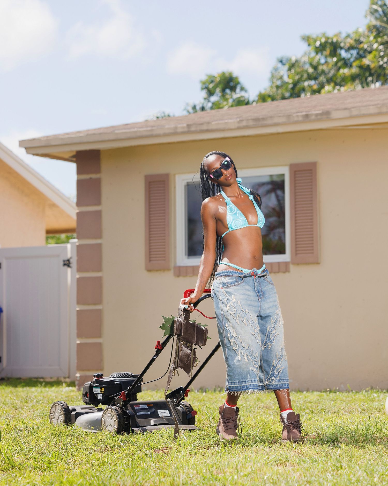 Woman in bikini top and jeans mowing lawn outside a suburban home on a sunny day.