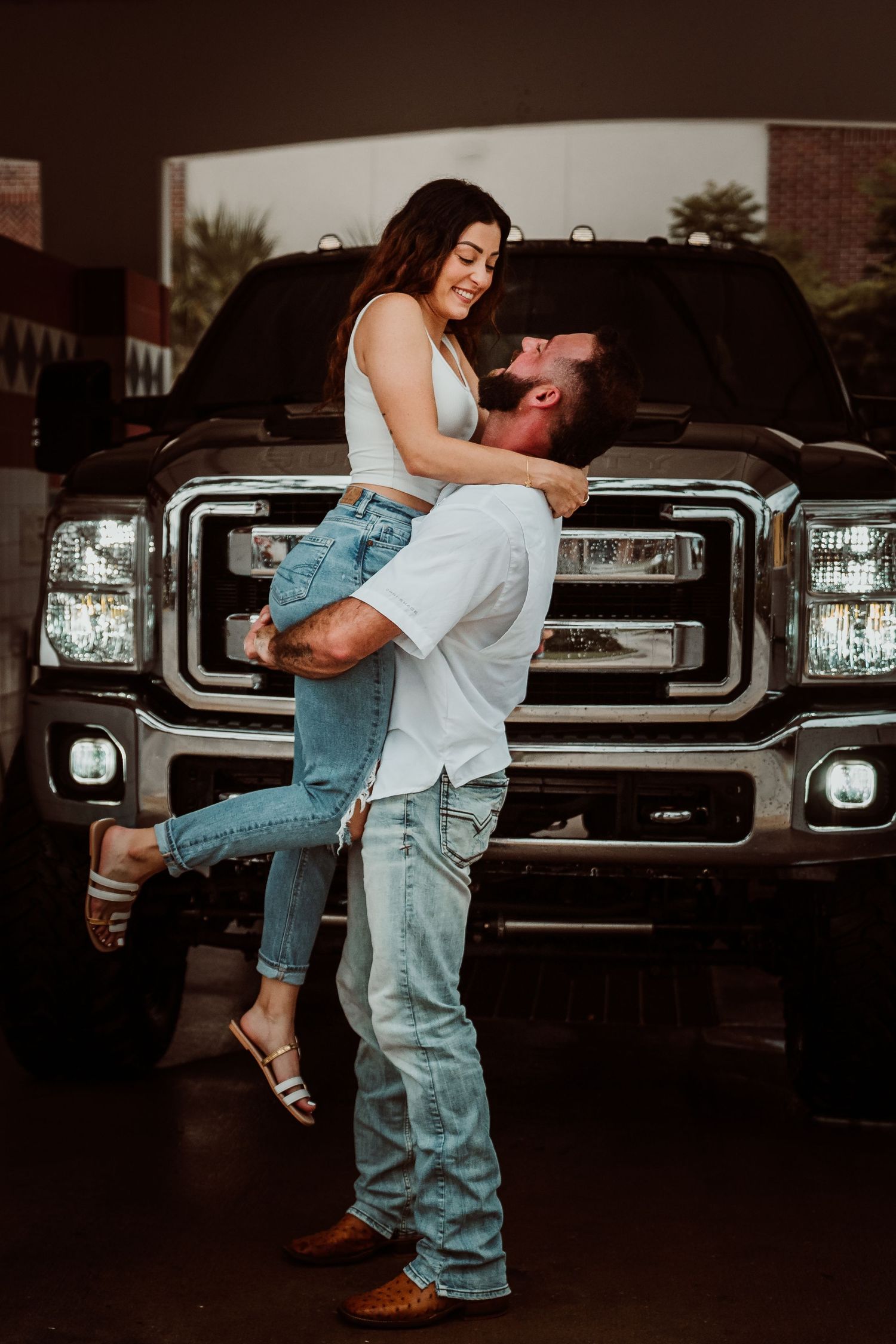 Man lifting woman in front of black Ford truck headlights in romantic engagement photo.
