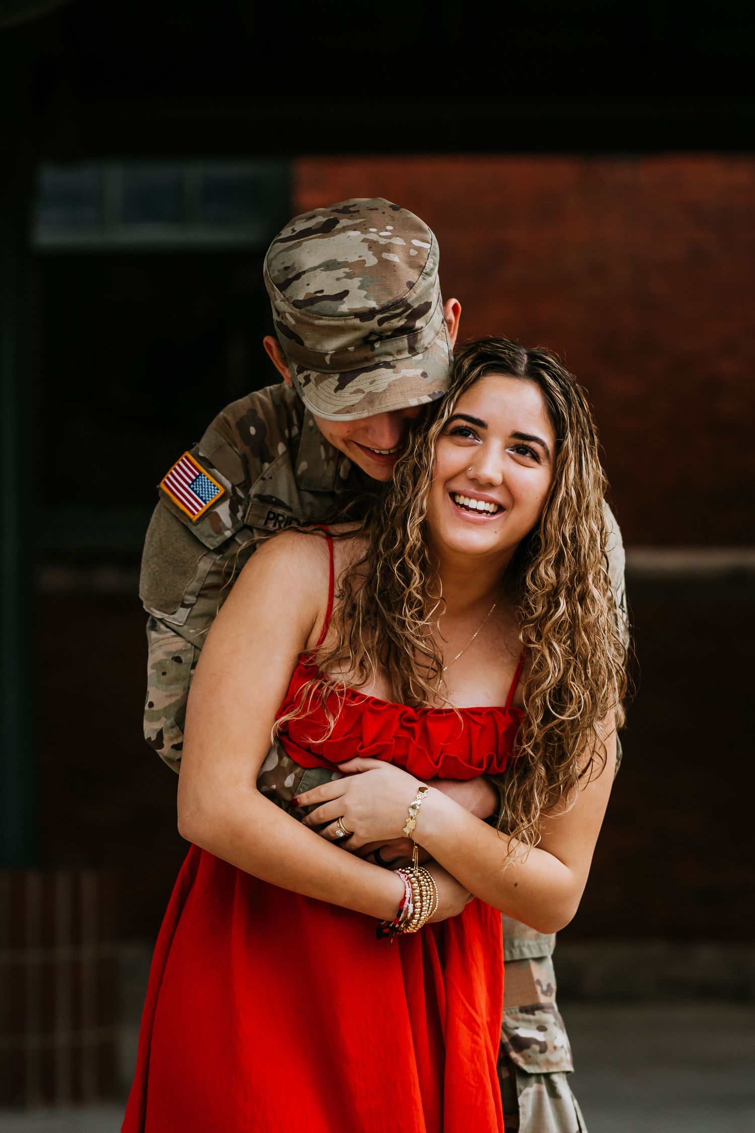 Smiling woman in red dress embraced from behind by US Army soldier in camouflage uniform.