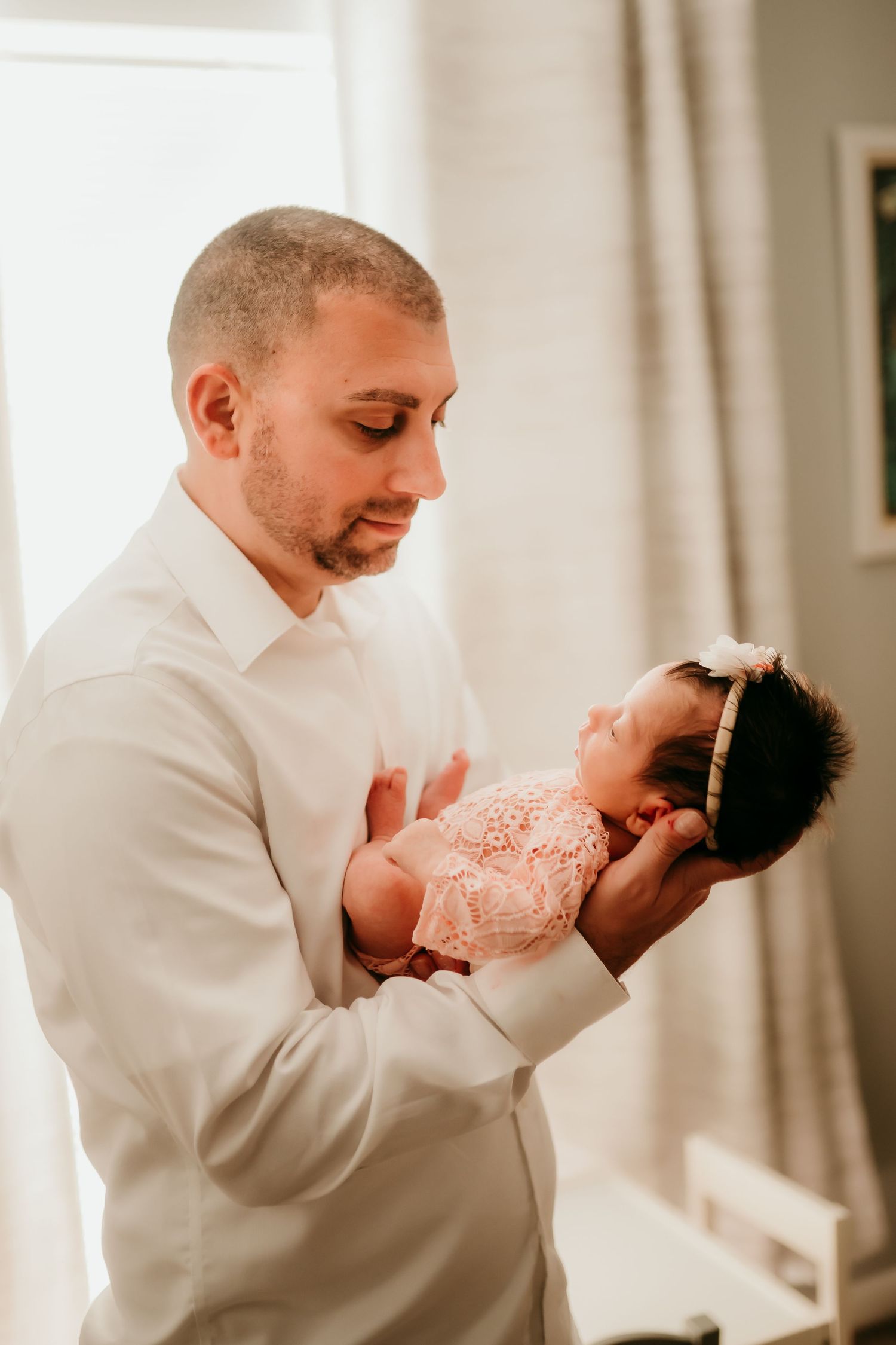 Father in white shirt tenderly holding his sleeping newborn baby girl near a bright window.