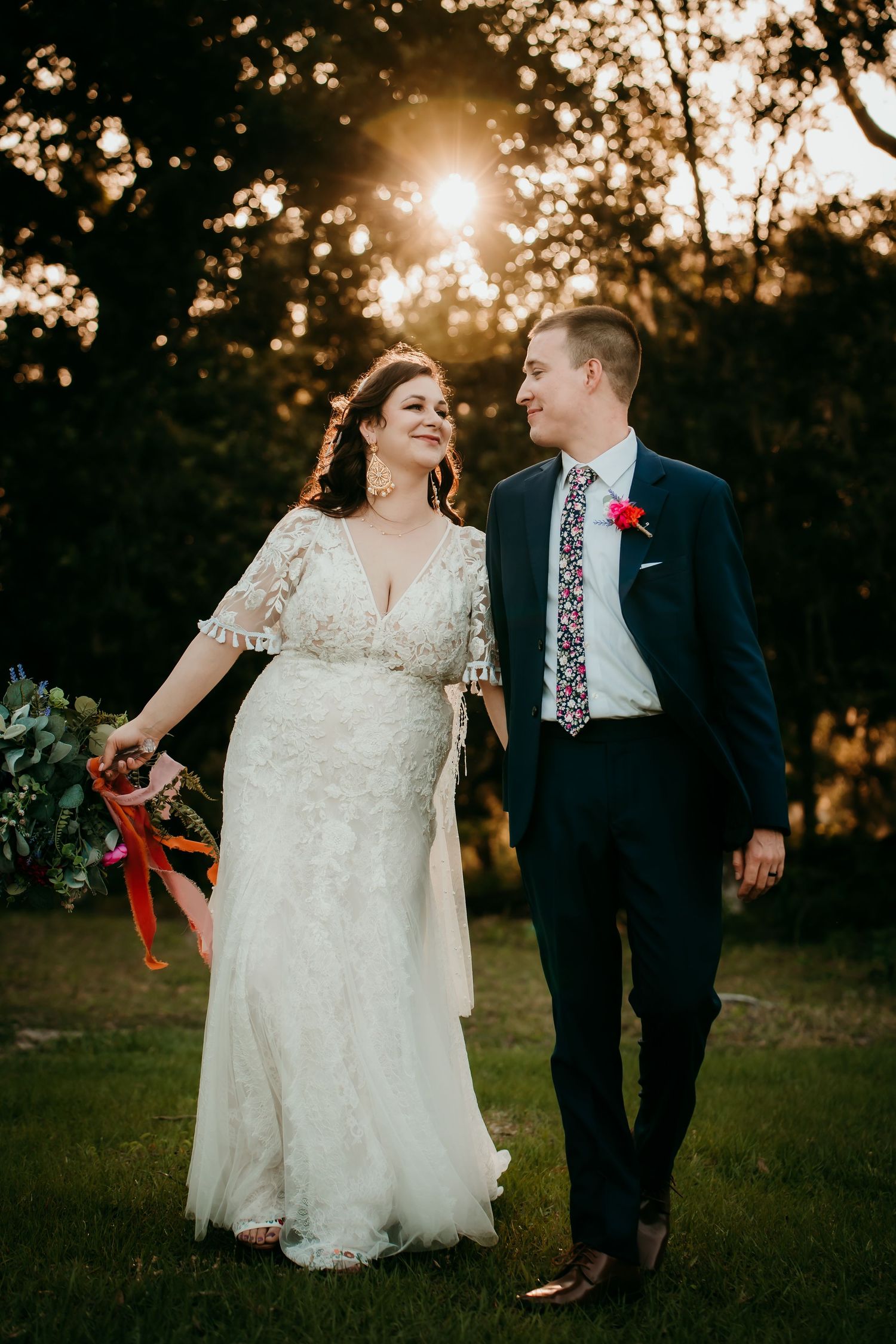 A bride and groom walk together at golden hour, smiling, as sunlight filters through trees behind them.