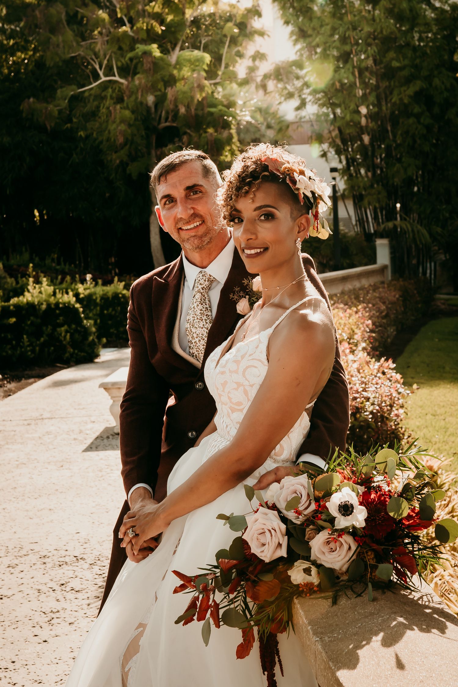 Smiling bride holding bouquet of red and pink flowers poses with groom in dark suit outdoors.