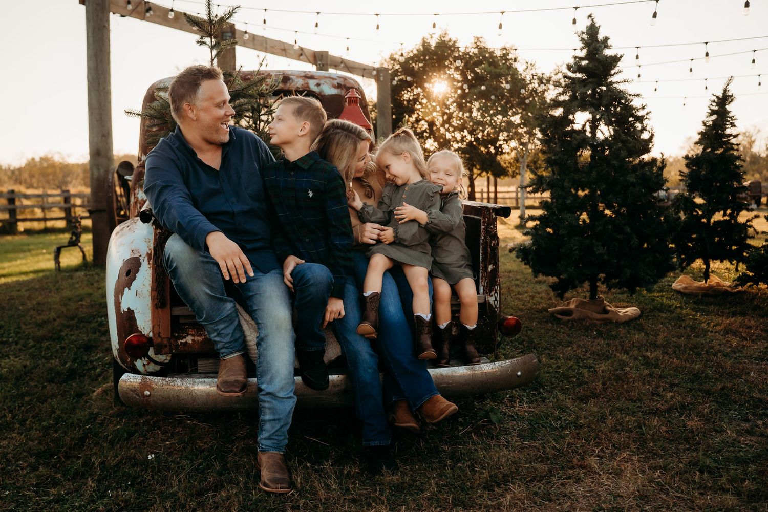 Happy family of five sitting on vintage red truck tailgate at sunset near Christmas trees.