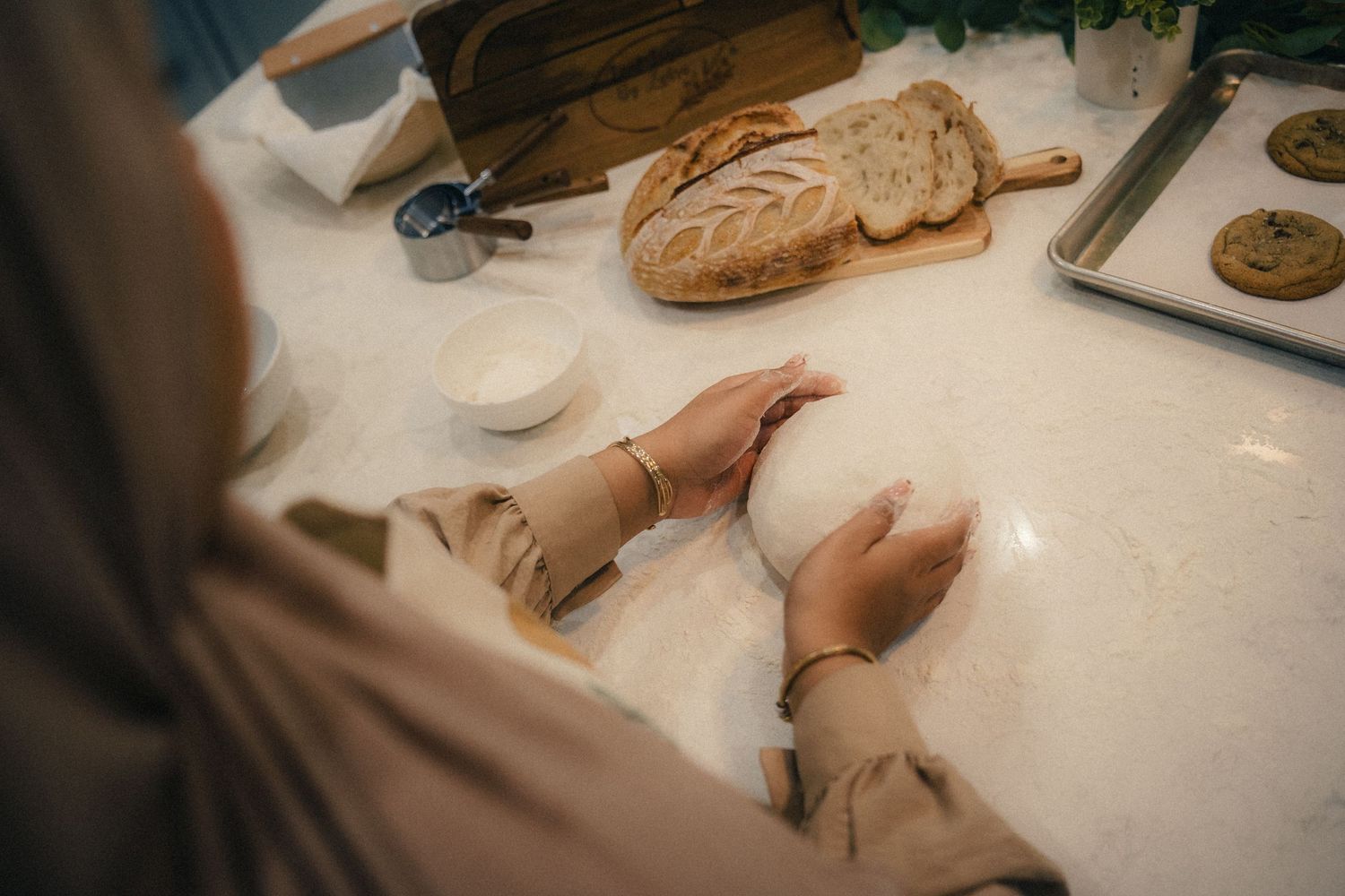 Person kneading dough on marble countertop with rustic bread loaf on wooden cutting board in bright kitchen.