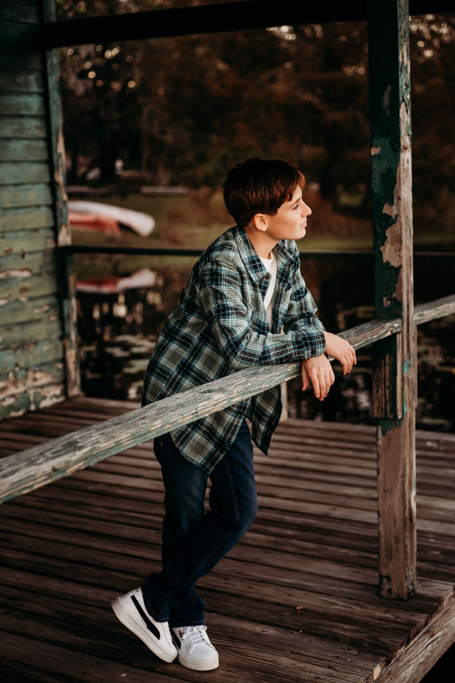 A young boy in a plaid shirt leans on a wooden railing of a rustic porch, gazing thoughtfully into the distance.