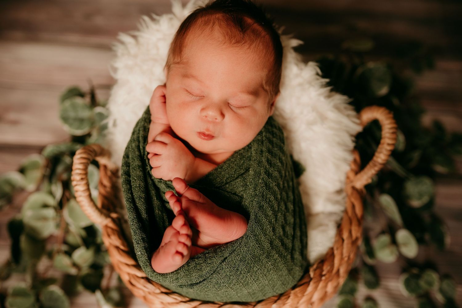 Sleeping newborn wrapped in green knit swaddle nestled in wicker basket with eucalyptus.
