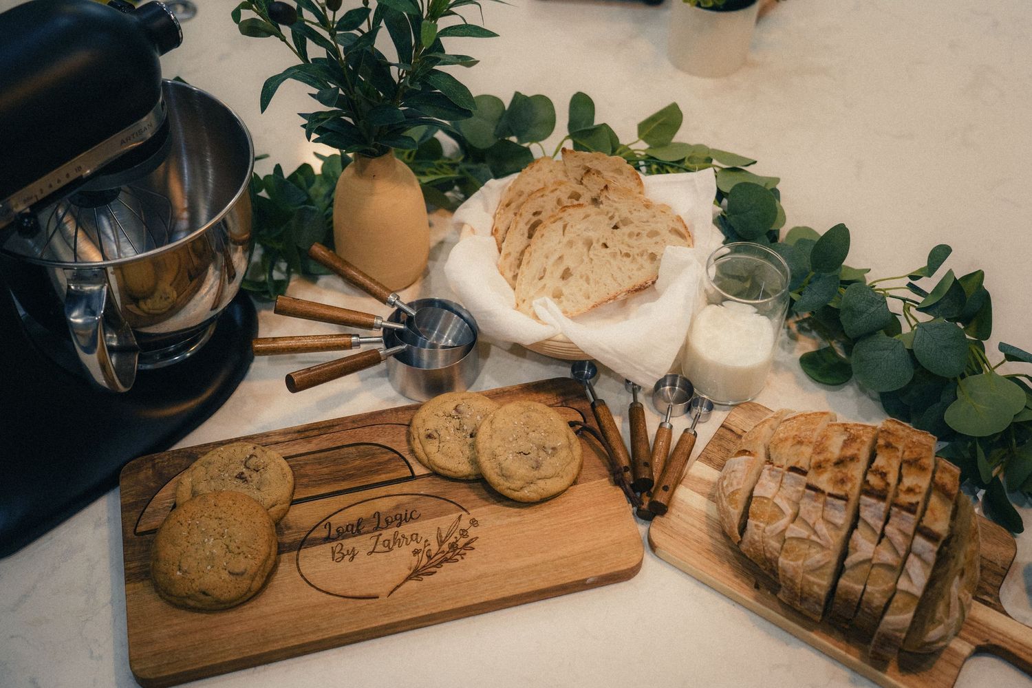 Wooden cutting boards with sliced bread, cookies, and baked goods displayed on a white marble countertop with greenery.