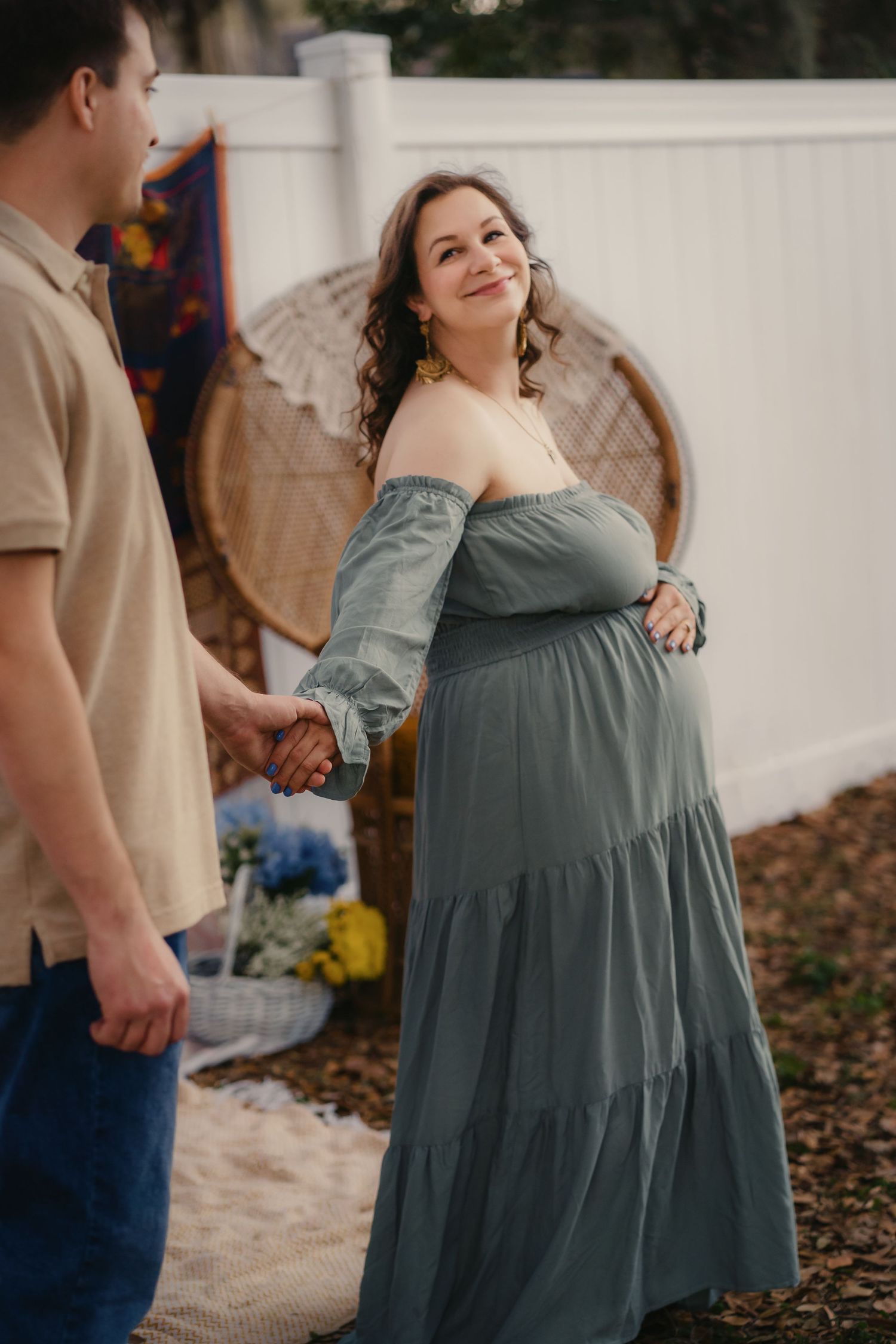 Pregnant woman in sage green off-shoulder dress holds hands with partner outdoors during maternity photo session.