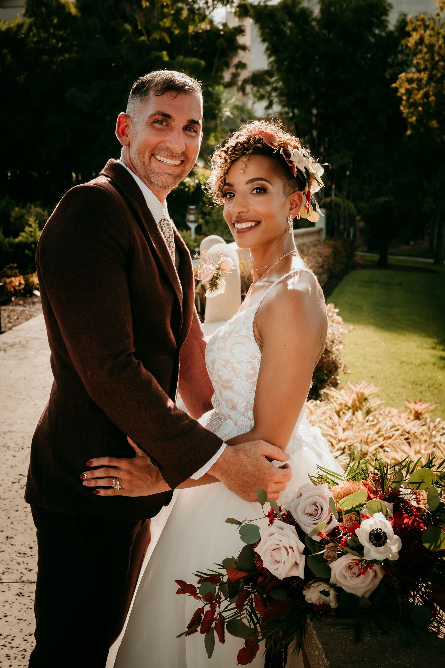 Smiling bride and groom pose outdoors in golden light, bride holding colorful bouquet of roses and anemones in white wedding dress.
