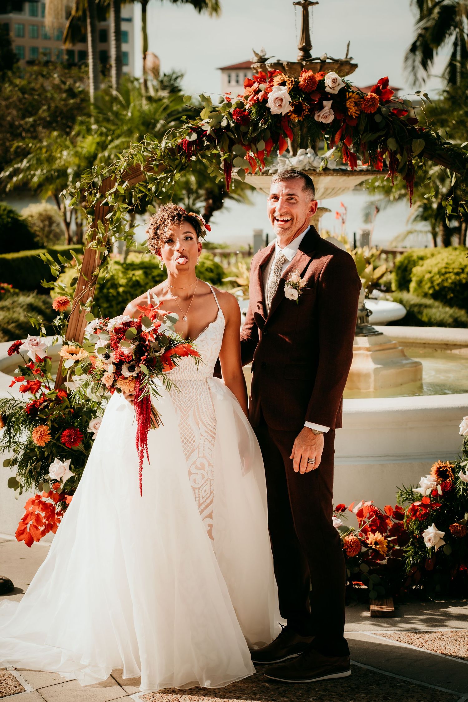 Bride and groom smiling at outdoor wedding ceremony with red floral arch and fountain backdrop.