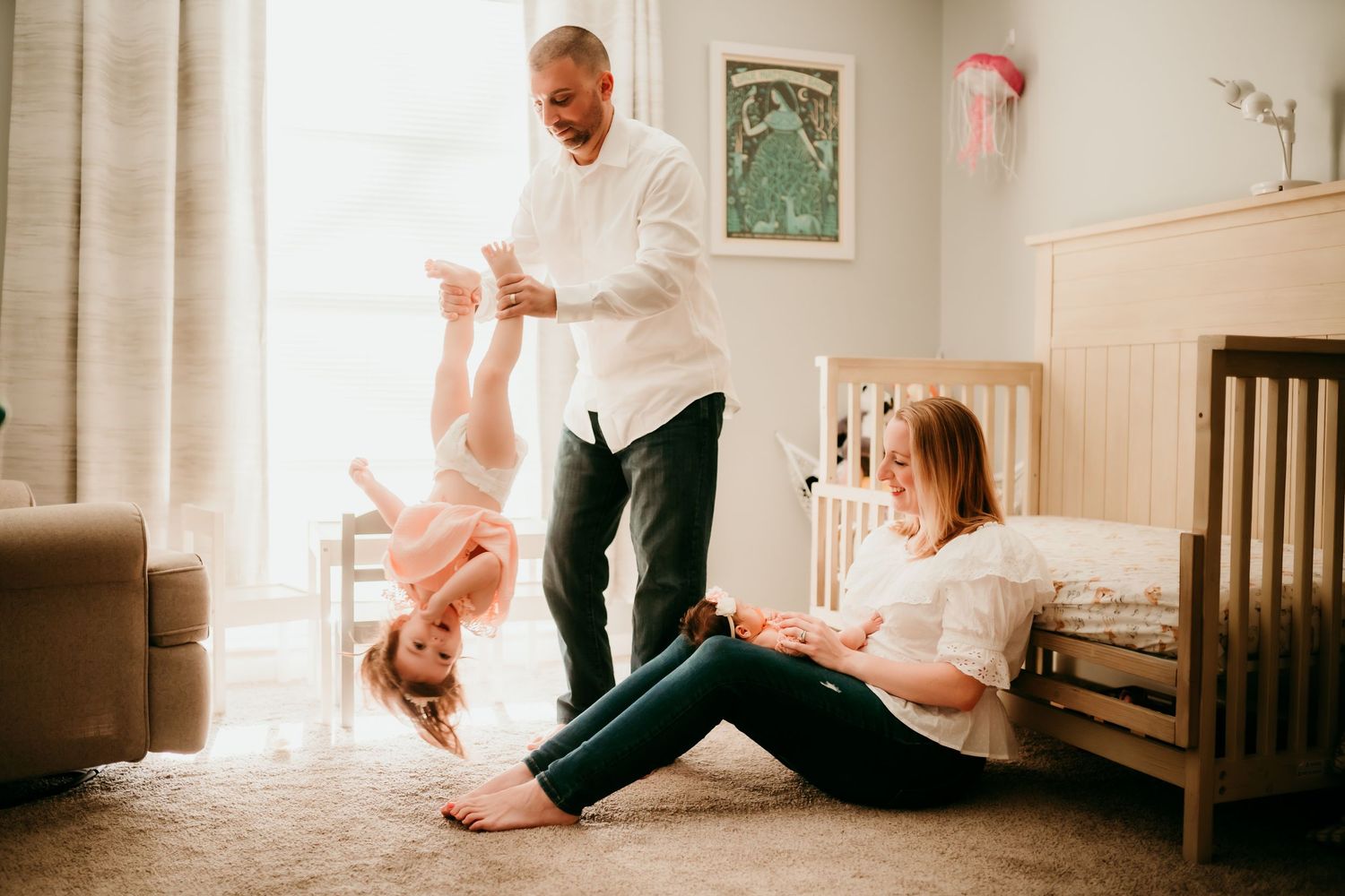 Family playing in nursery with father holding toddler upside down while mother sits smiling.