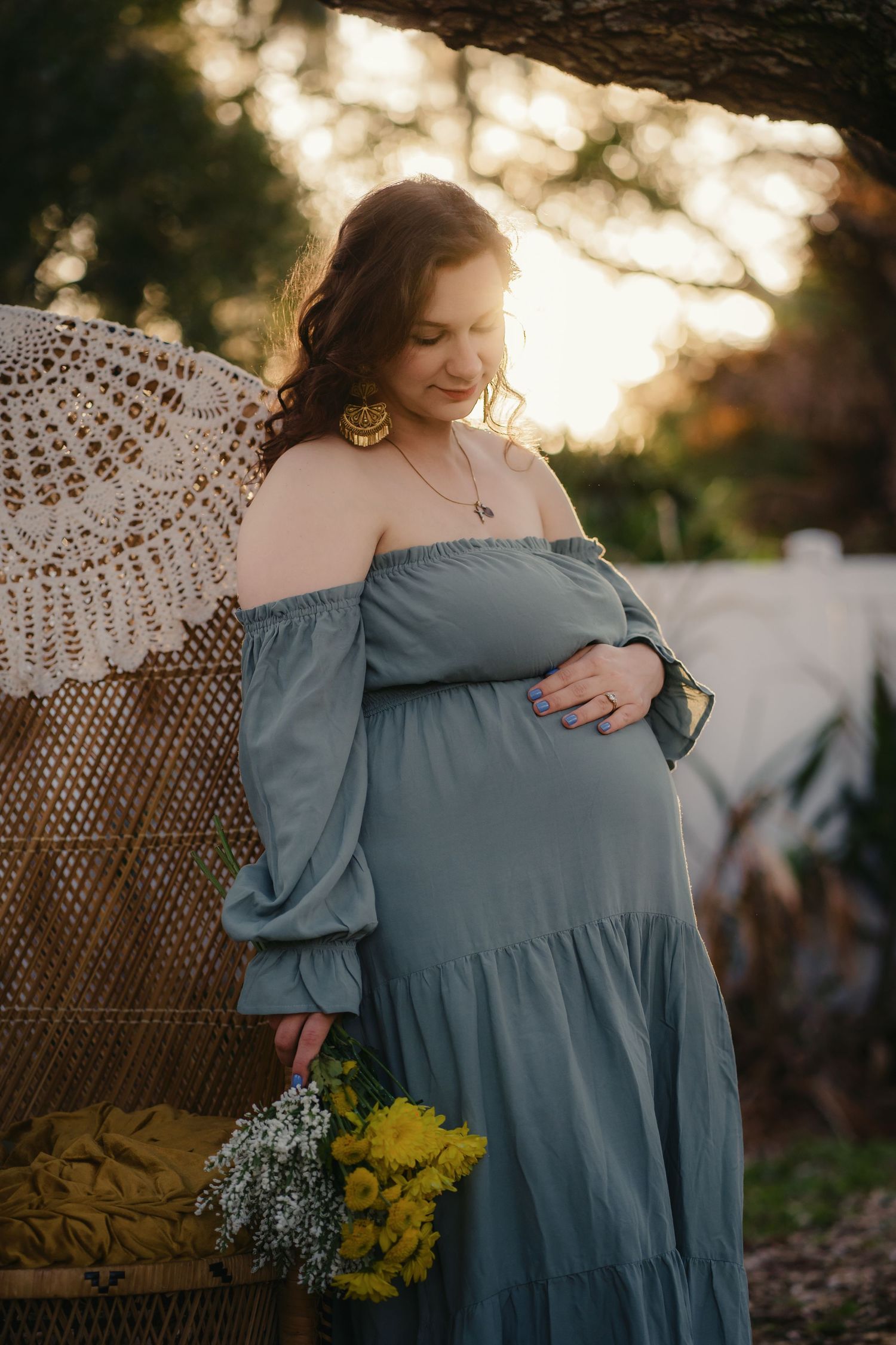 Pregnant woman in teal off-shoulder dress holding yellow flowers at golden hour outdoors.