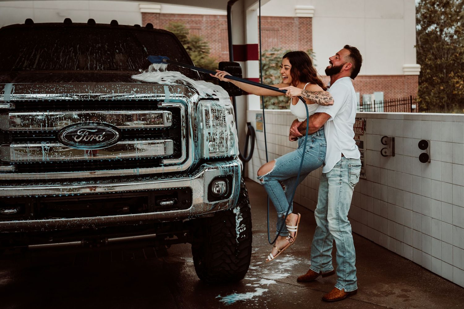 Playful couple washing a soapy black Ford truck together at an outdoor car wash.