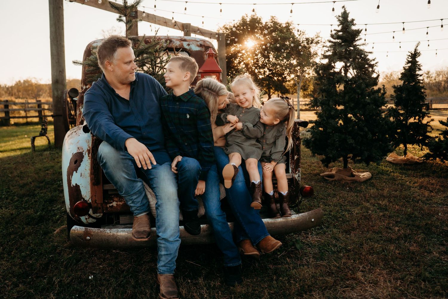 Father laughing with three children seated on vintage truck tailgate at golden hour near Christmas trees.