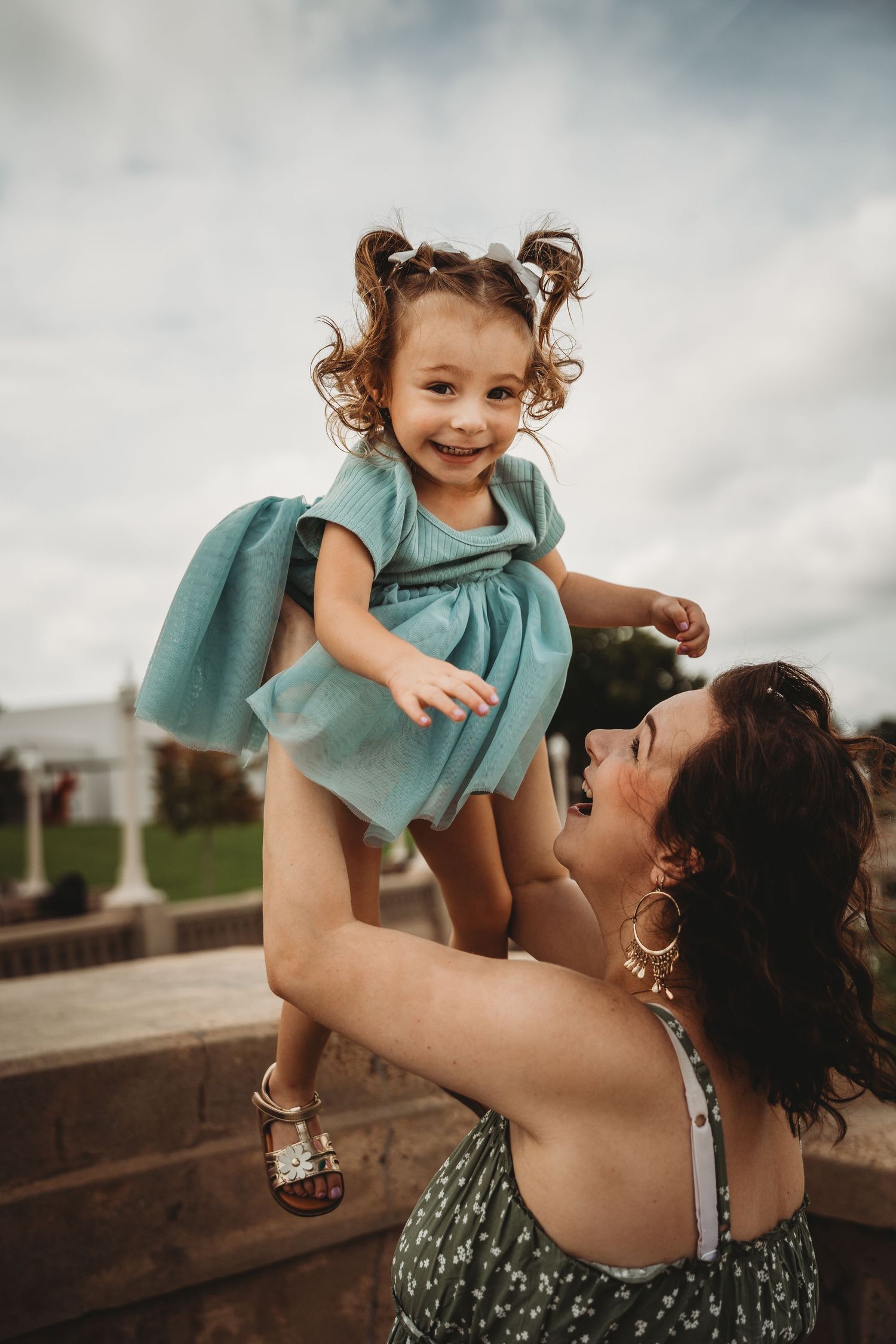 Mother lifting her happy toddler girl in a teal dress against a cloudy sky.