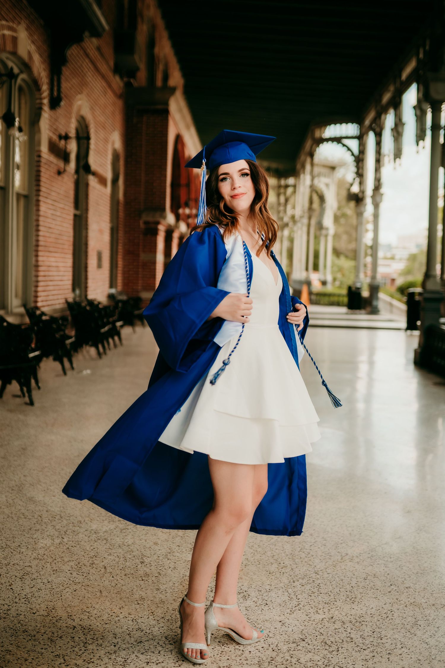Young woman in blue graduation cap and gown over white dress posing in elegant university hallway.