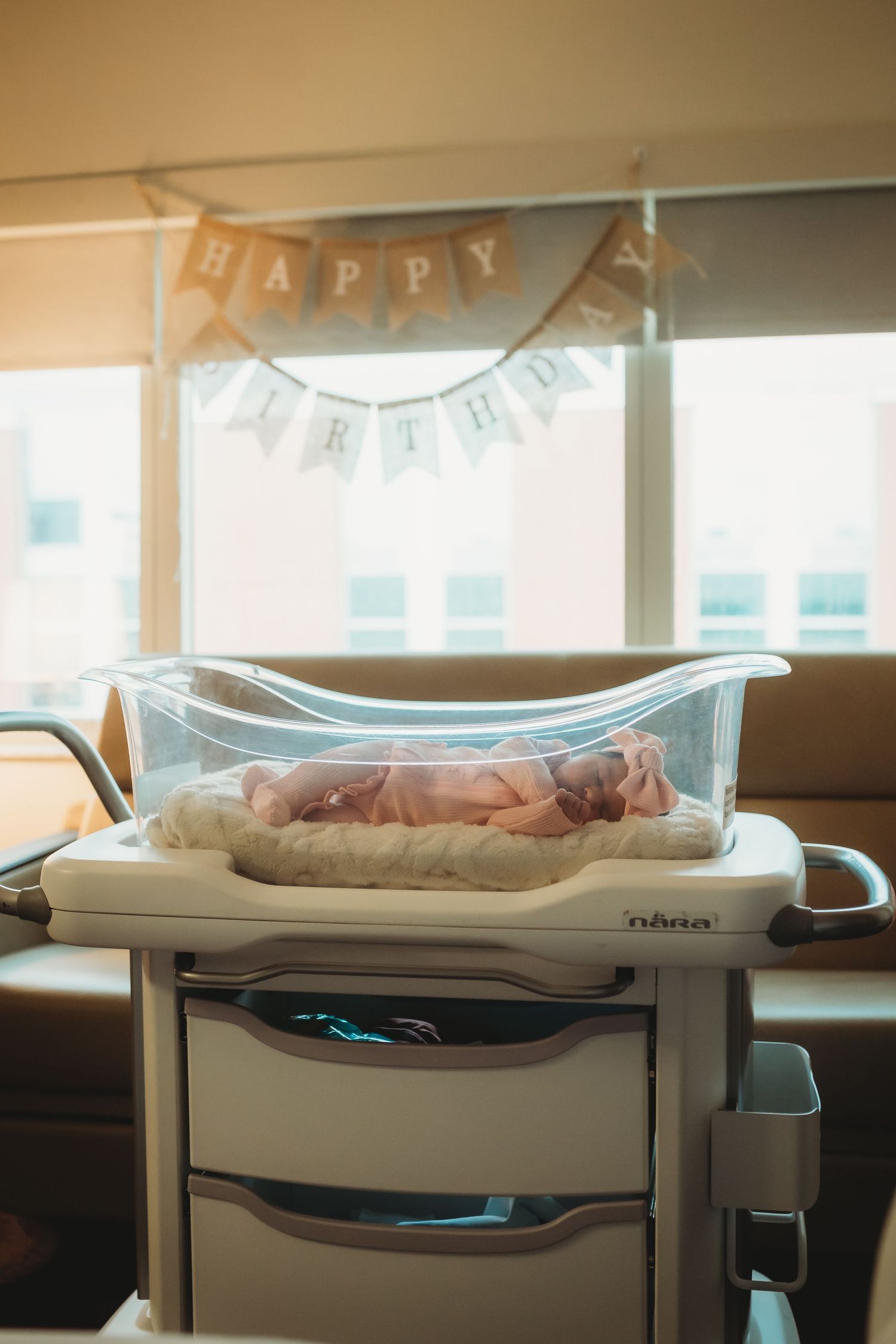 Sleeping newborn baby in hospital bassinet under Happy Birthday banner in warm light.