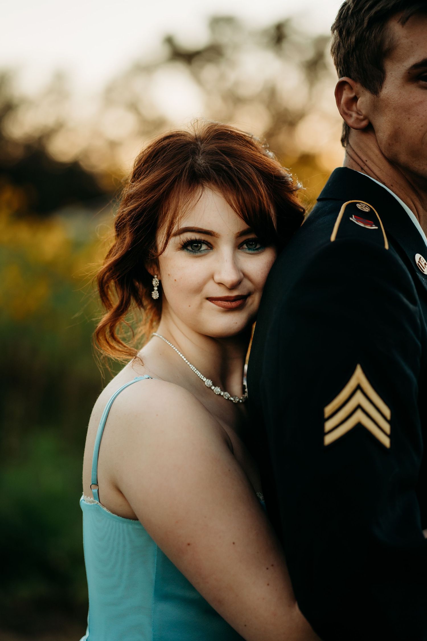 Young woman with auburn hair in blue gown leans against soldier's shoulder at golden hour.