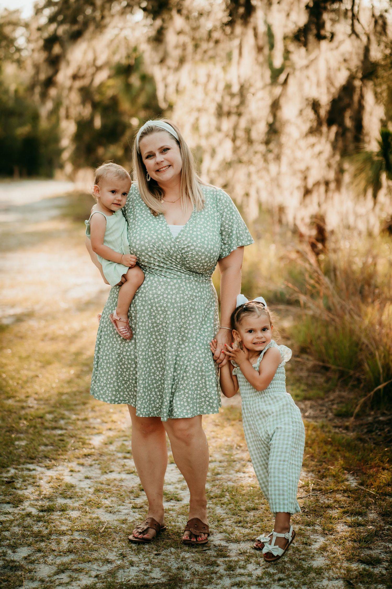 Mother in green floral dress holds toddler while older daughter clings to her hand outdoors.