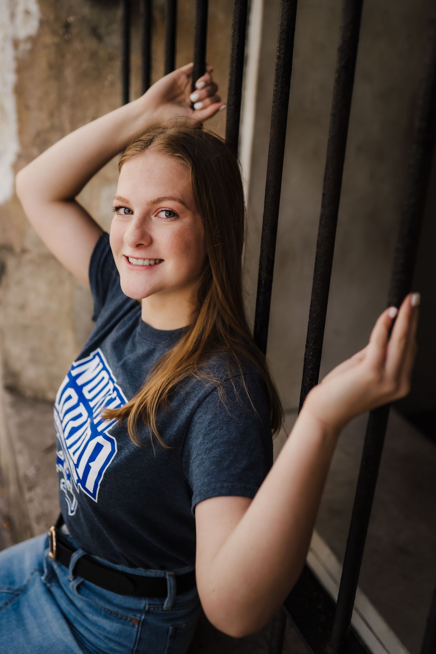 Smiling young woman in a navy graphic t-shirt and jeans leaning against black metal railings outdoors.