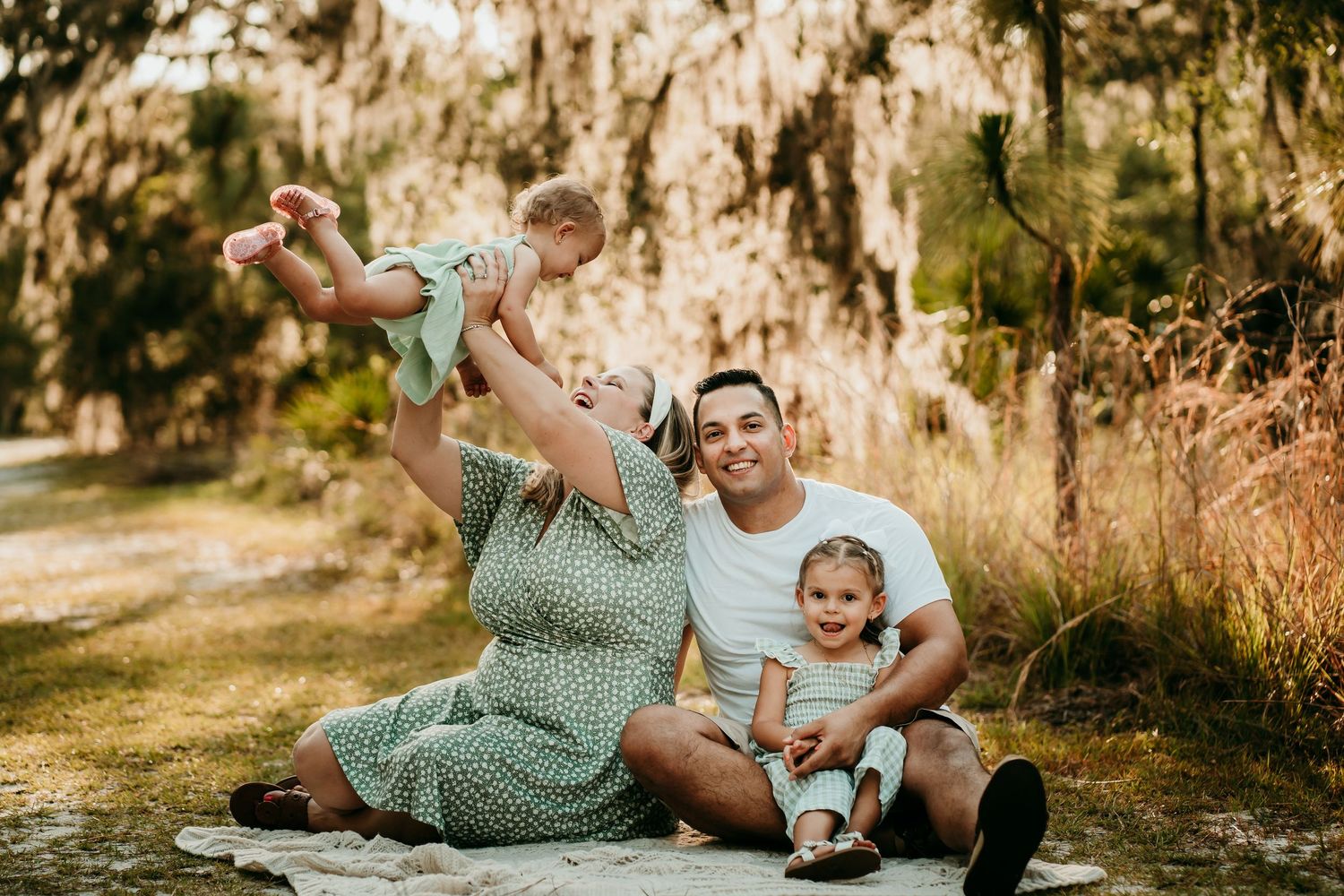 Joyful family outdoors as father lifts smiling baby while mother and toddler sit nearby.