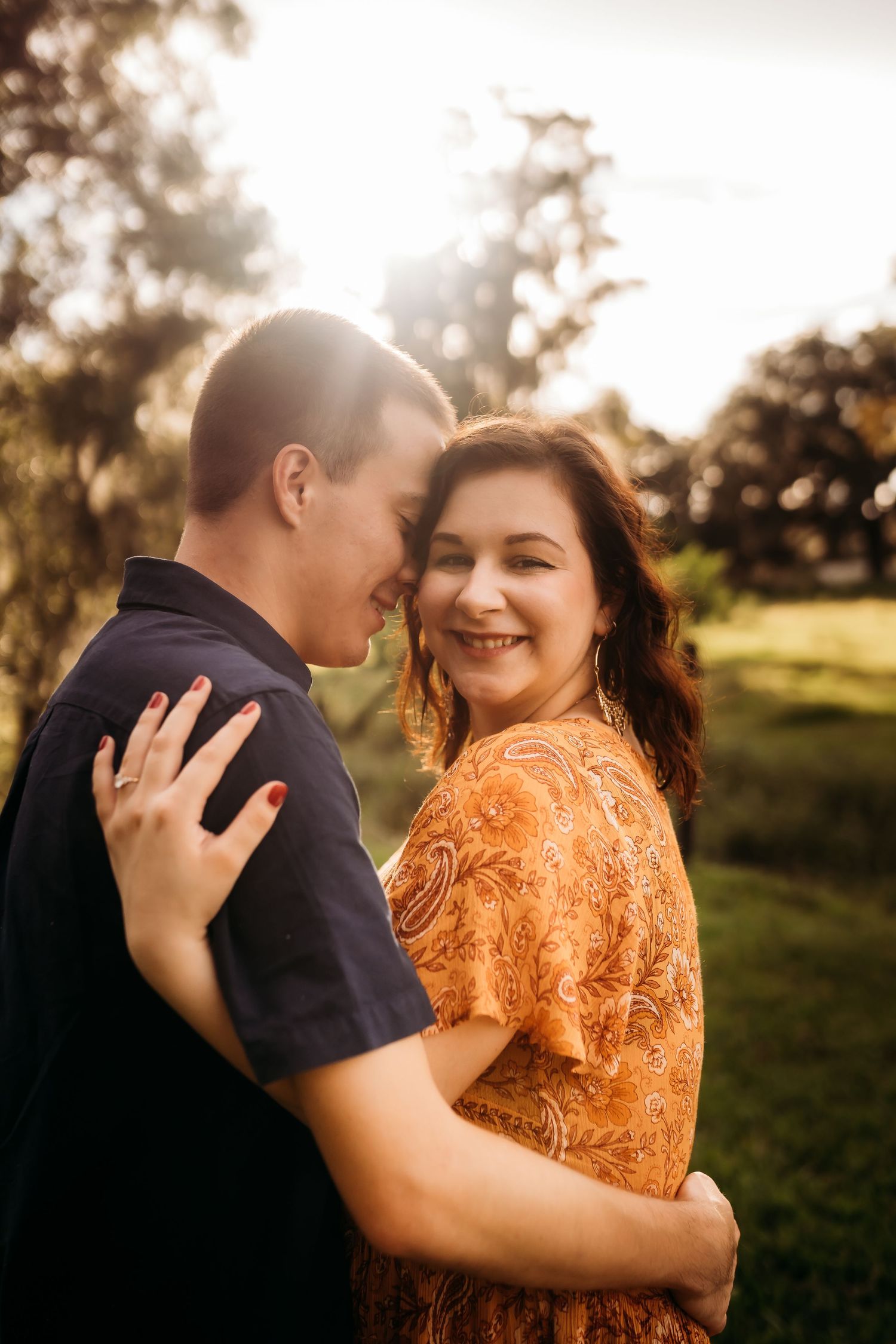 A smiling woman in an orange dress embraces a man in a navy shirt during a golden sunset in a park.