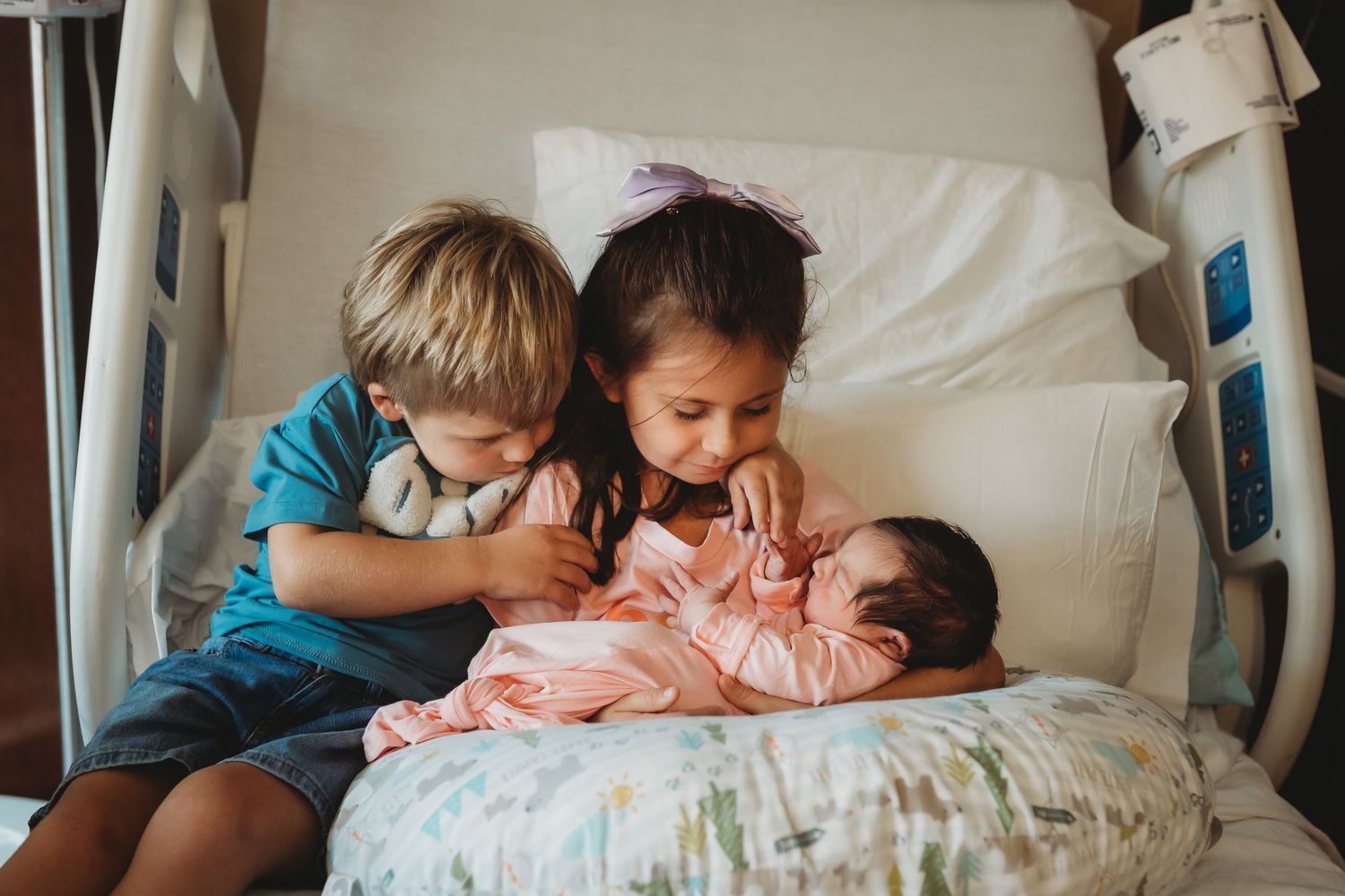 Two young siblings gently holding newborn baby sister on hospital bed.