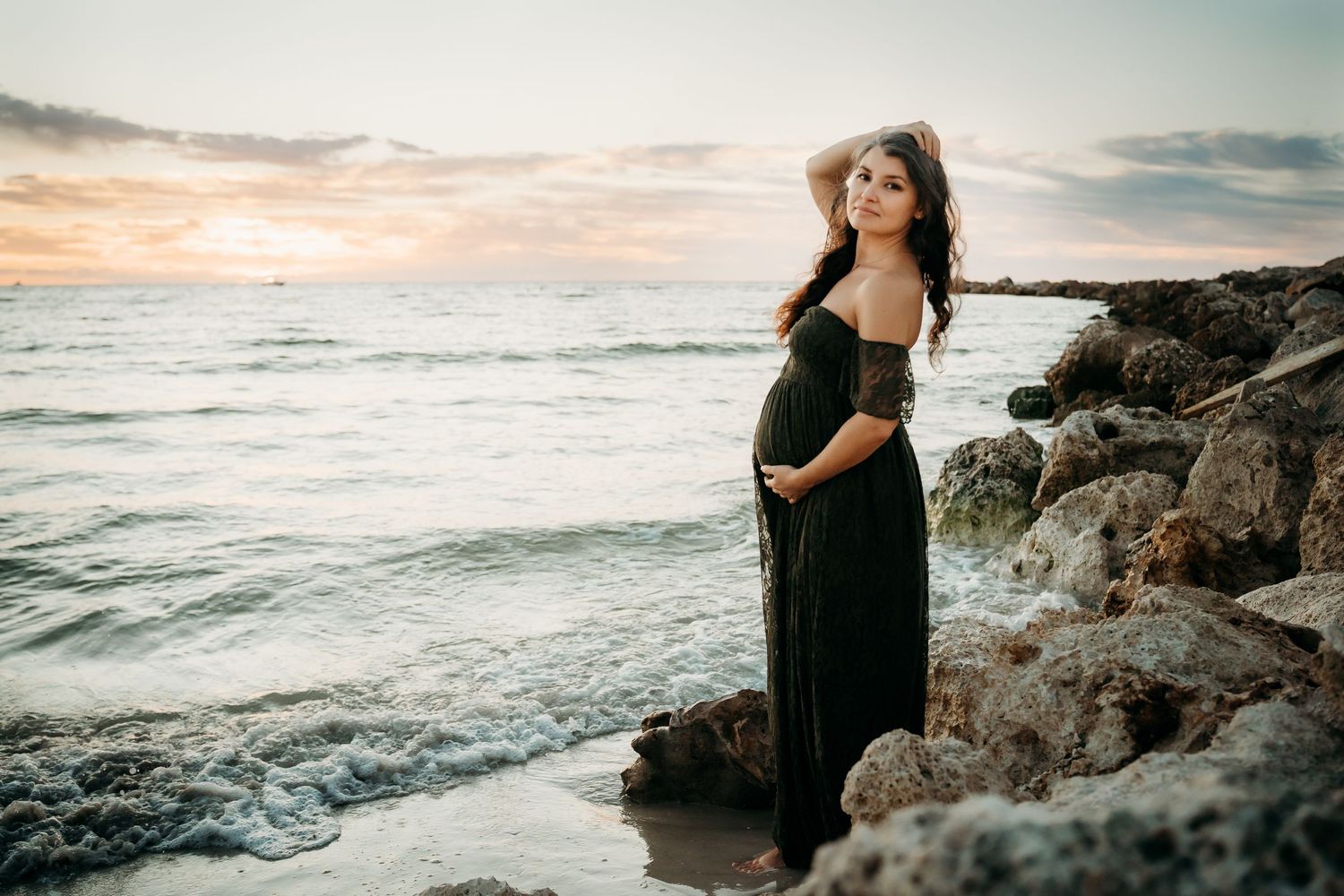 Pregnant woman in dark green off-shoulder gown poses on rocky beach at sunset.