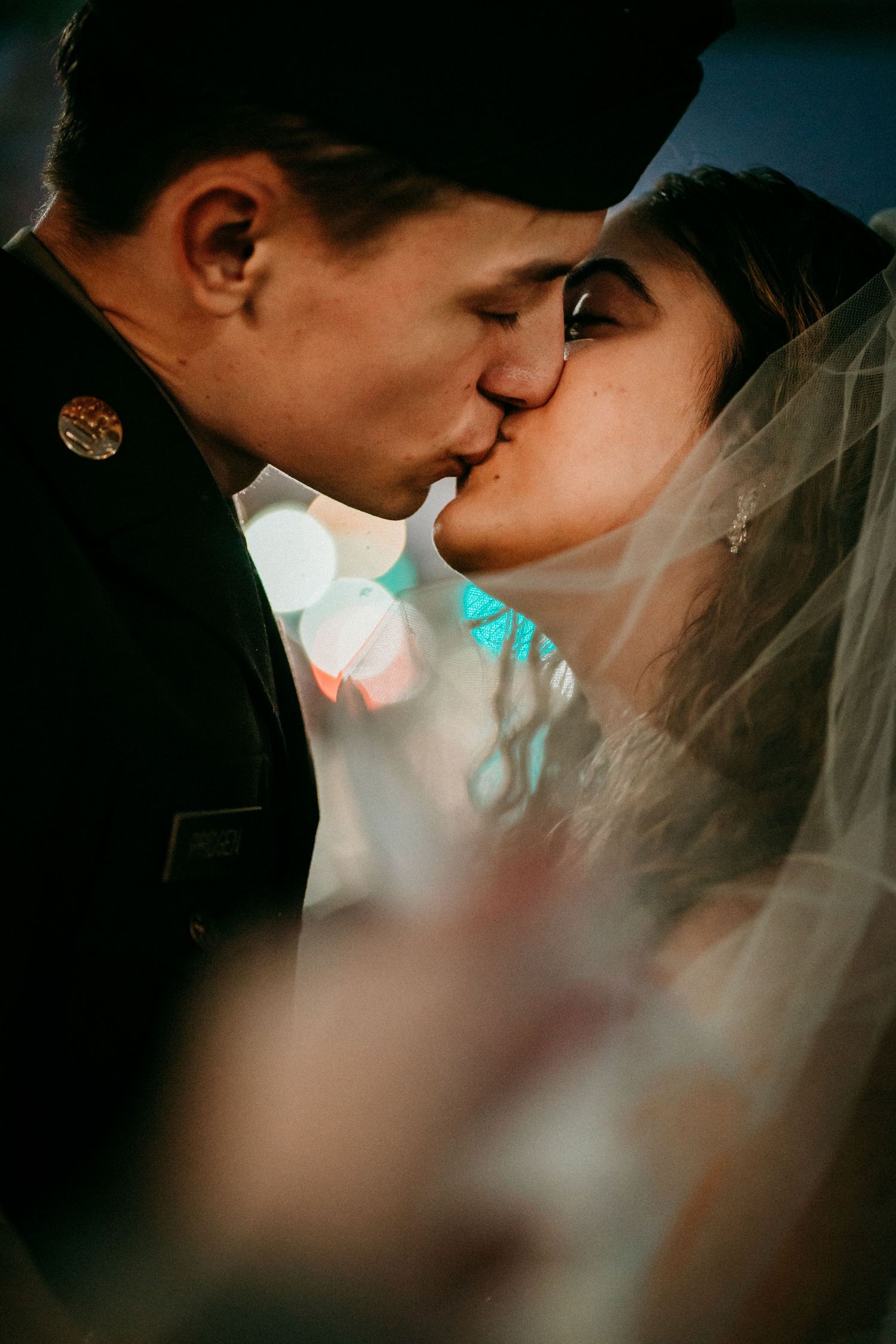 Bride and groom share romantic kiss under veil with colorful bokeh lights in background.
