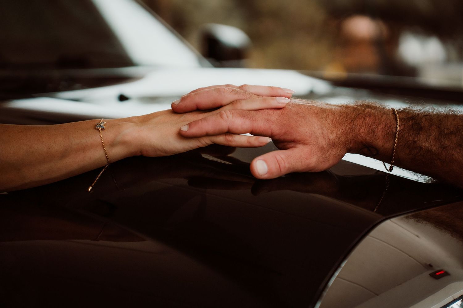 Close-up of couple holding hands gently on a dark car hood, wearing delicate bracelets.