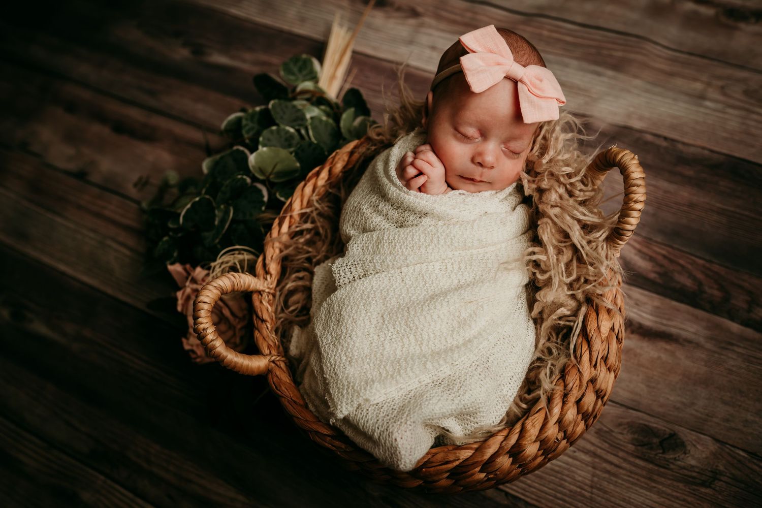 Newborn baby swaddled in cream wrap with pink bow sleeps in wicker basket with greenery.