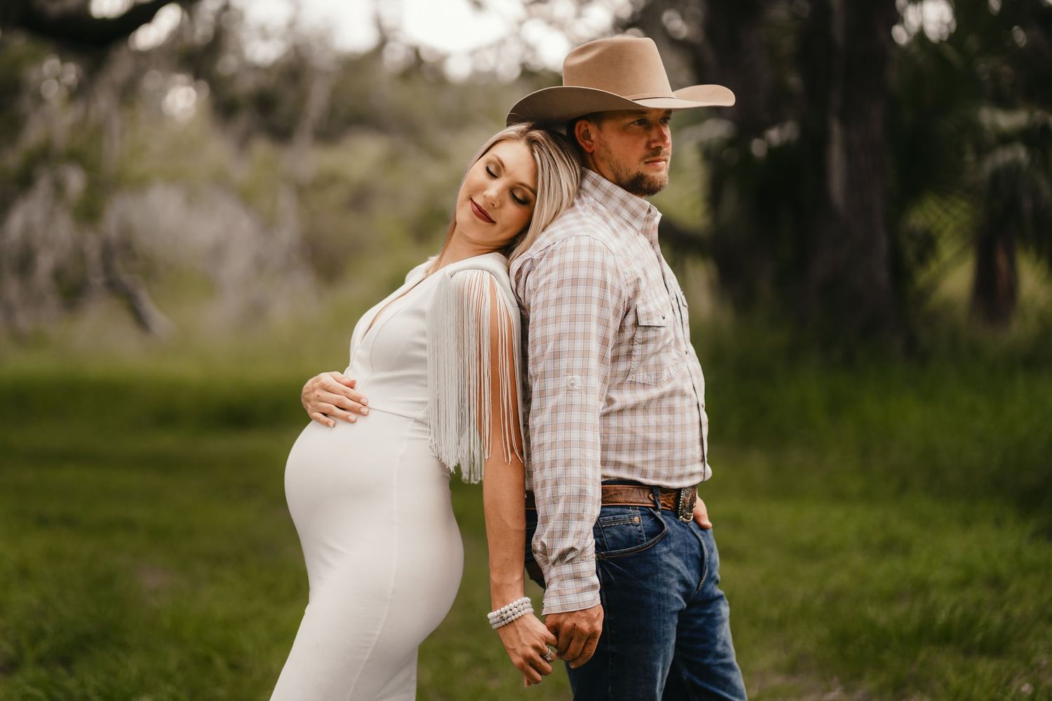 A pregnant woman in a white dress leans back-to-back with a man in a cowboy hat during a golden hour maternity shoot.