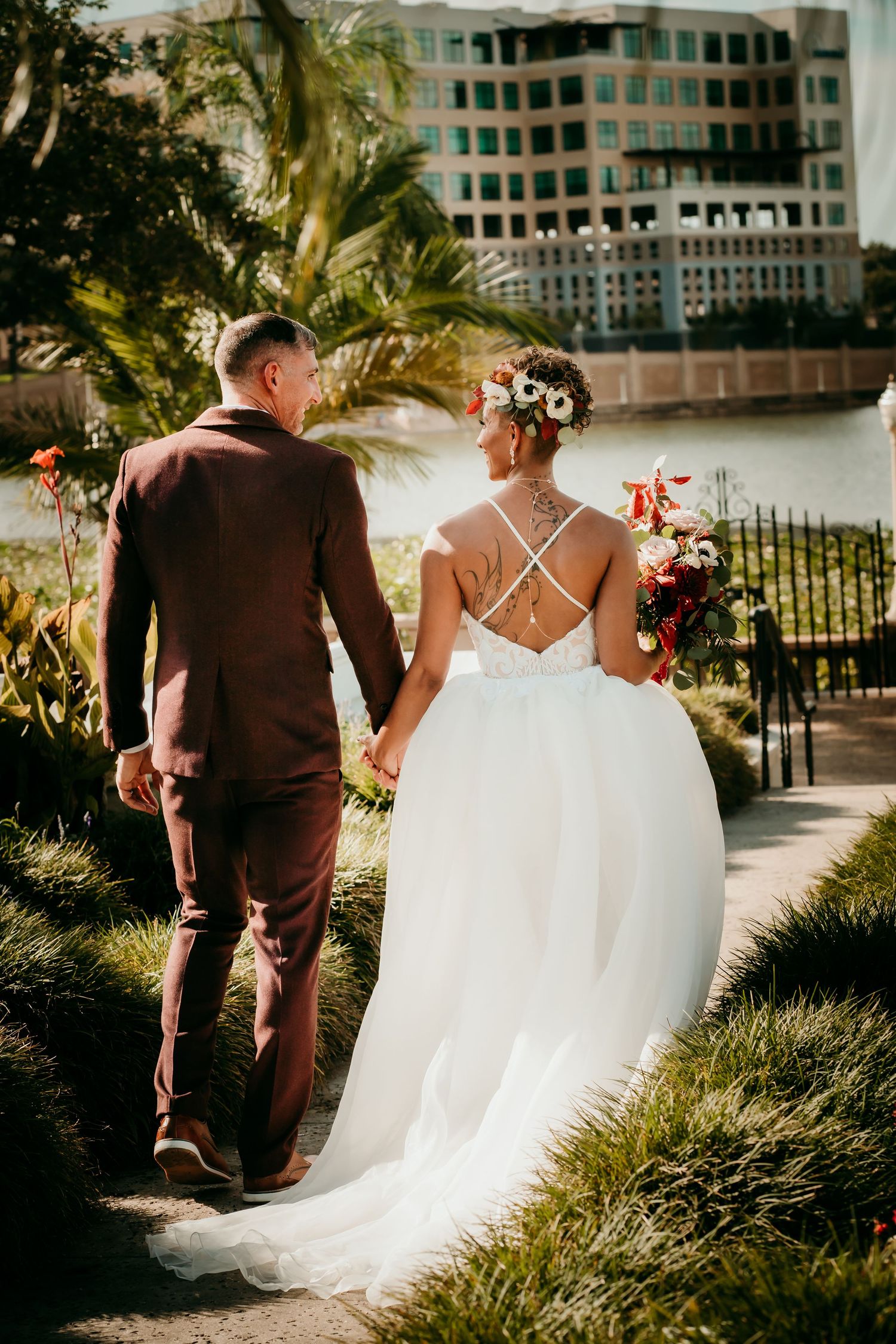 Bride and groom walking hand-in-hand, bride in white gown with floral crown holding bouquet, groom in burgundy suit.