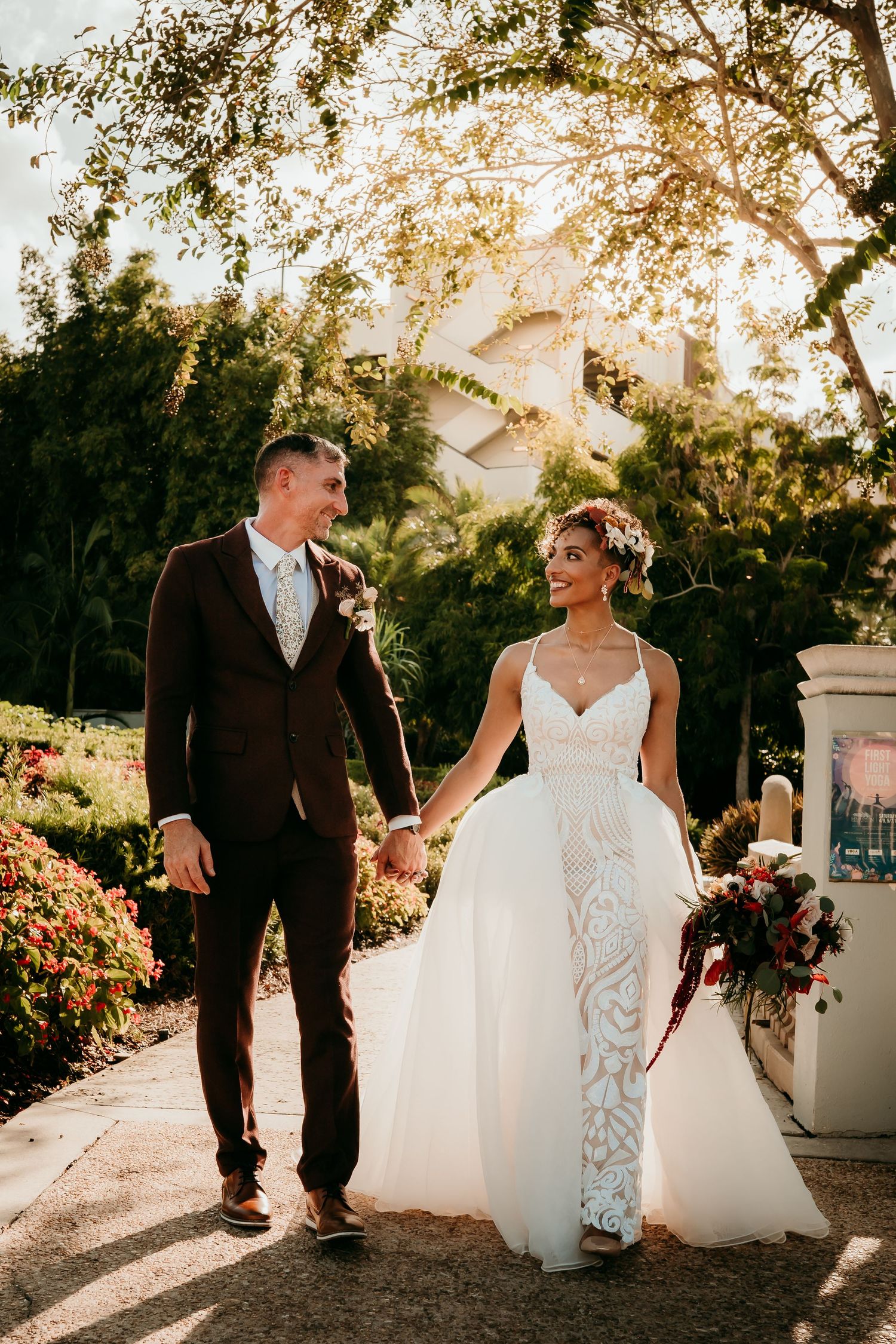 Bride and groom walk hand in hand through a sunlit garden, she in lace gown, he in dark suit.